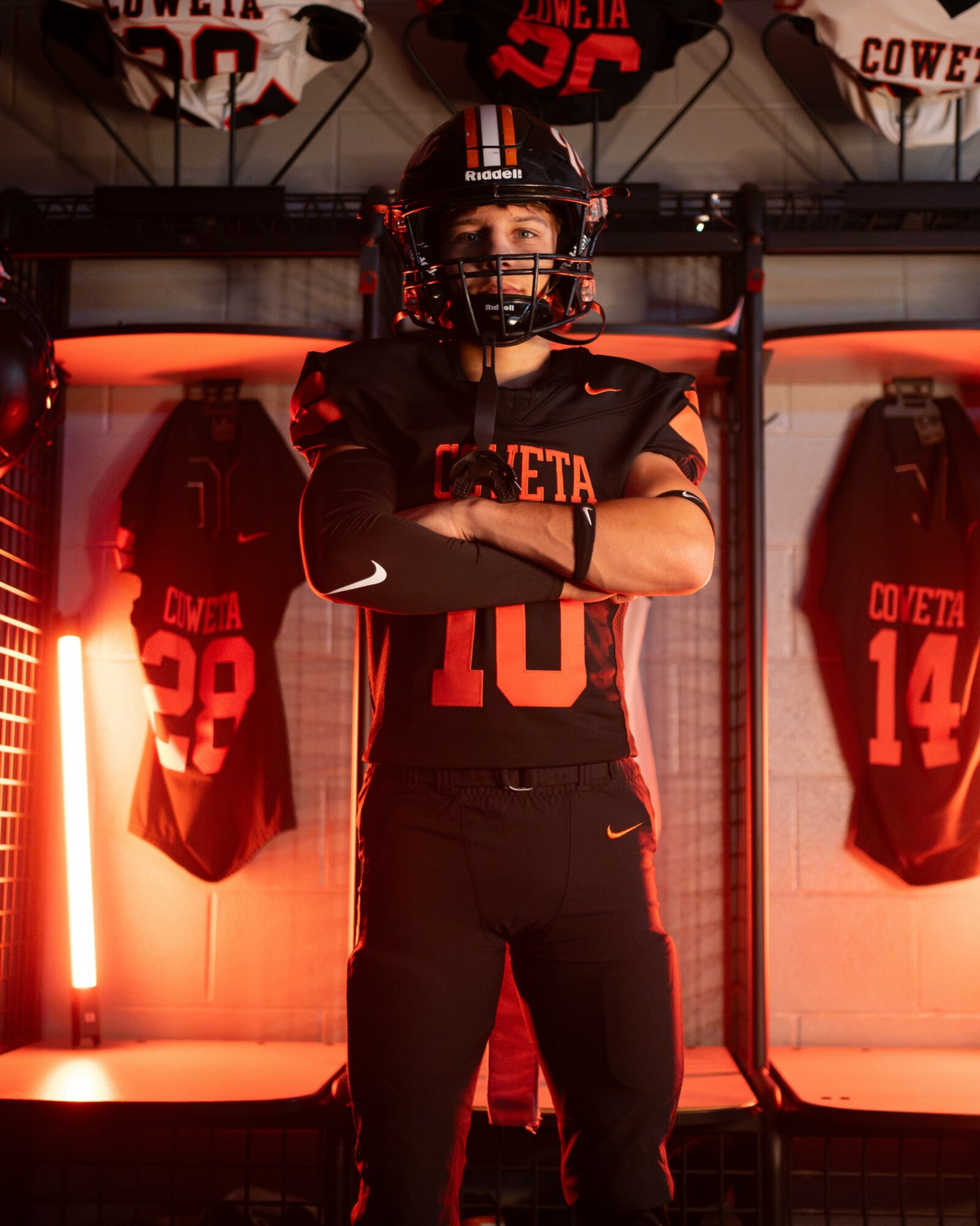 Person in football uniform with arms crossed, standing in front of jerseys and lockers, illuminated by red lighting.