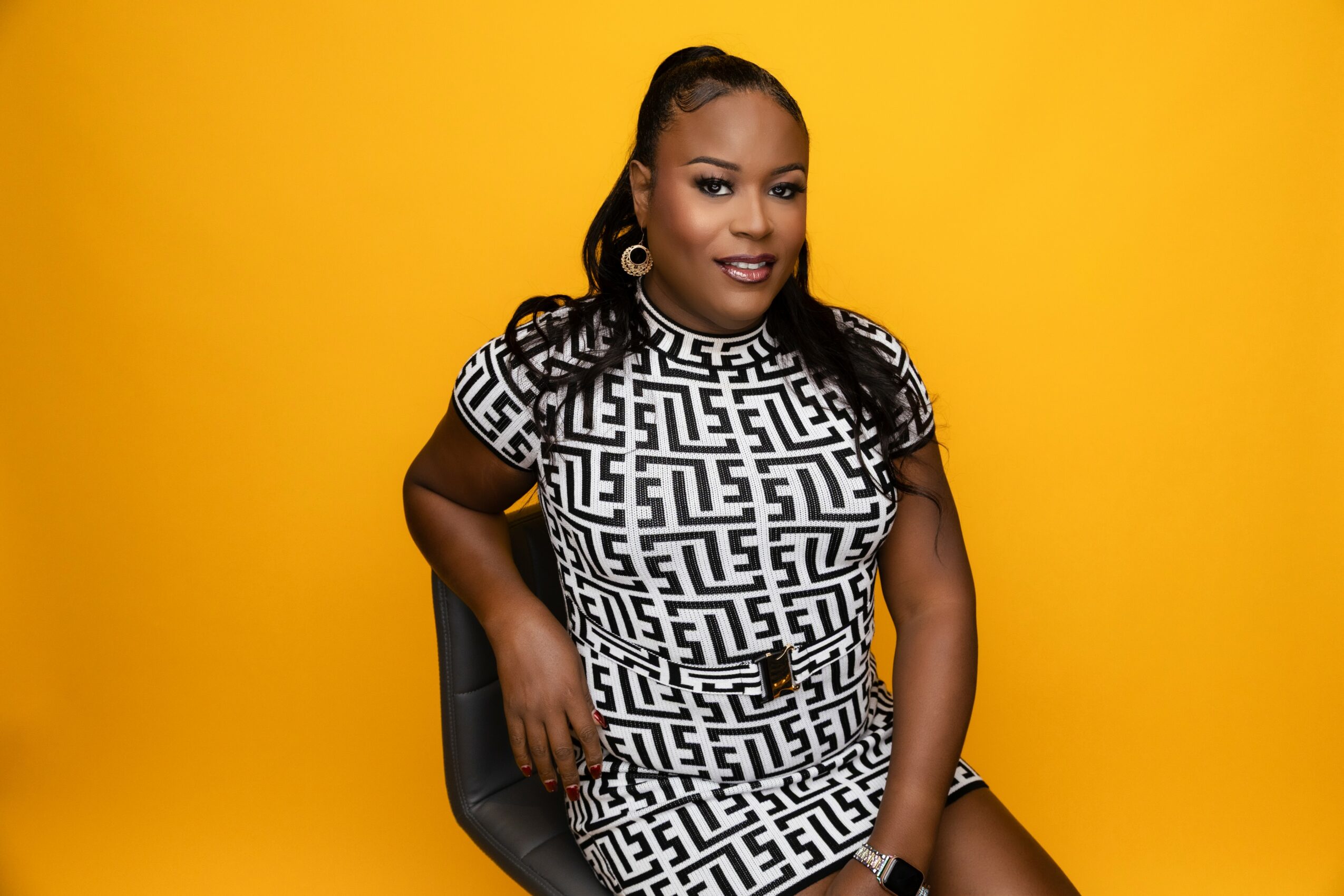Woman with long hair and earrings sitting against a yellow background, wearing a black and white patterned dress.