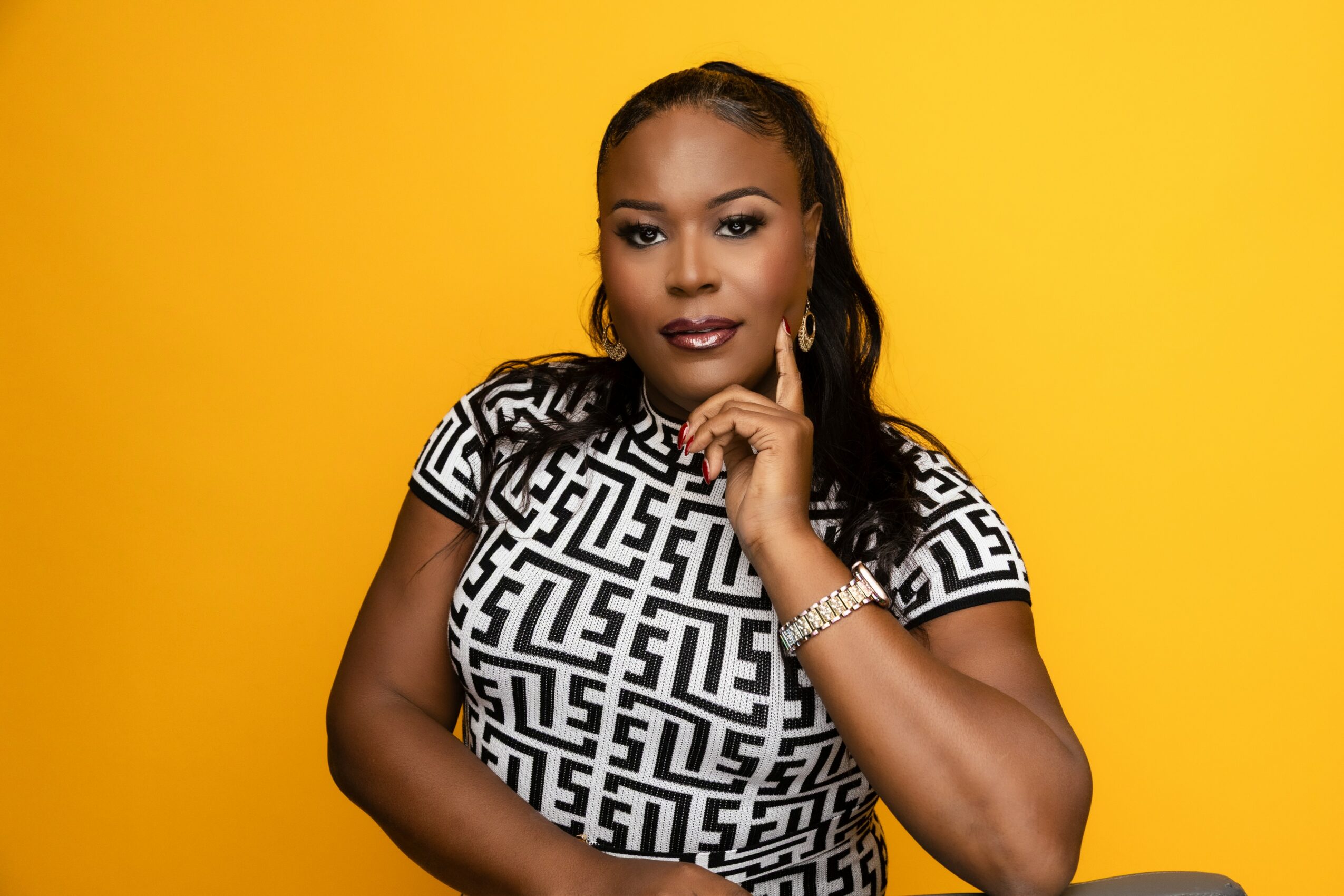 Woman with dark hair and makeup, wearing a patterned black and white top, poses against a yellow background.