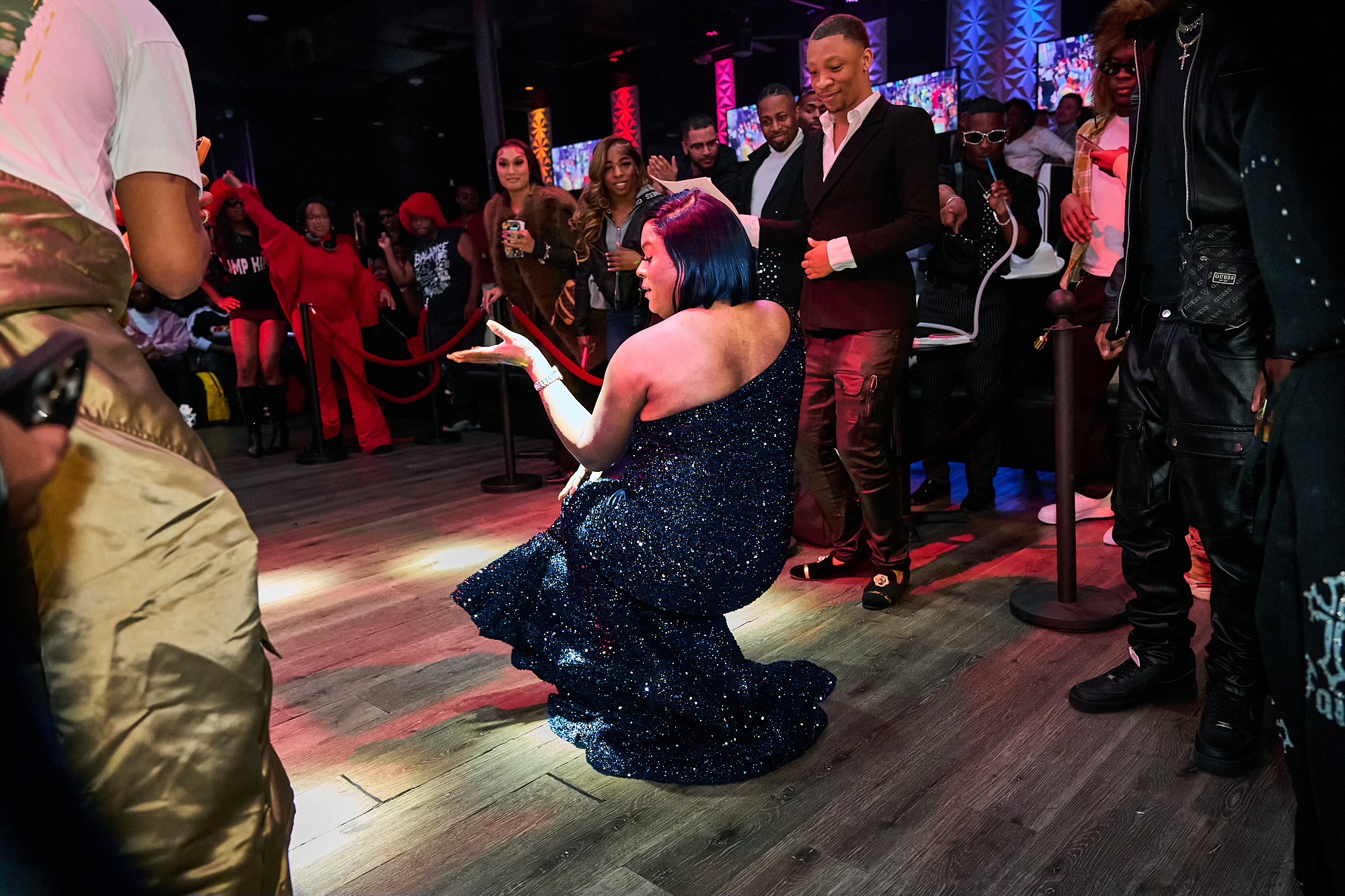 Person with short dark hair kneels on the dance floor, surrounded by people in a lively nightclub scene.