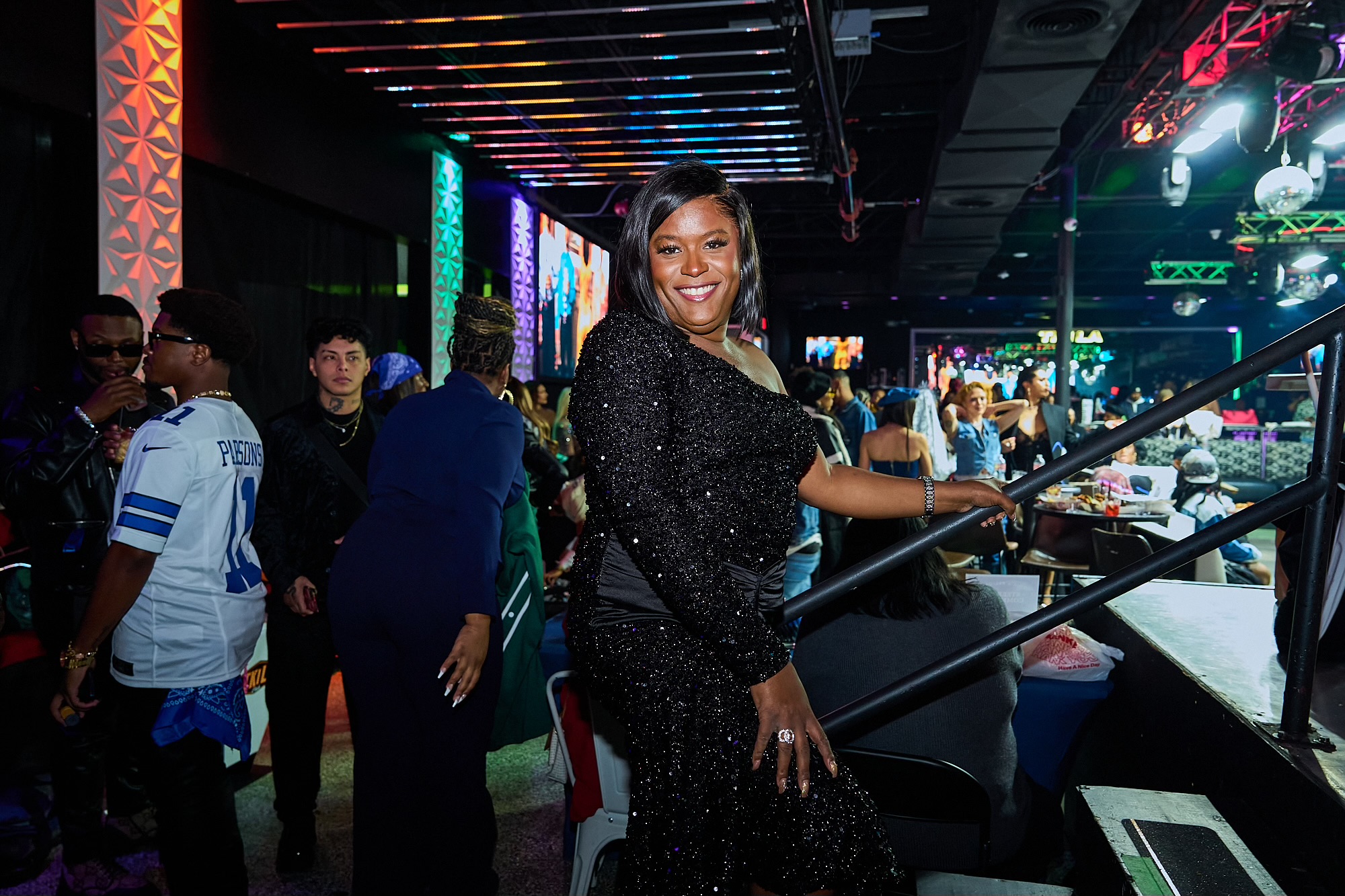Woman smiling and standing at a bar in a lively nightclub with colorful lights and people in the background.