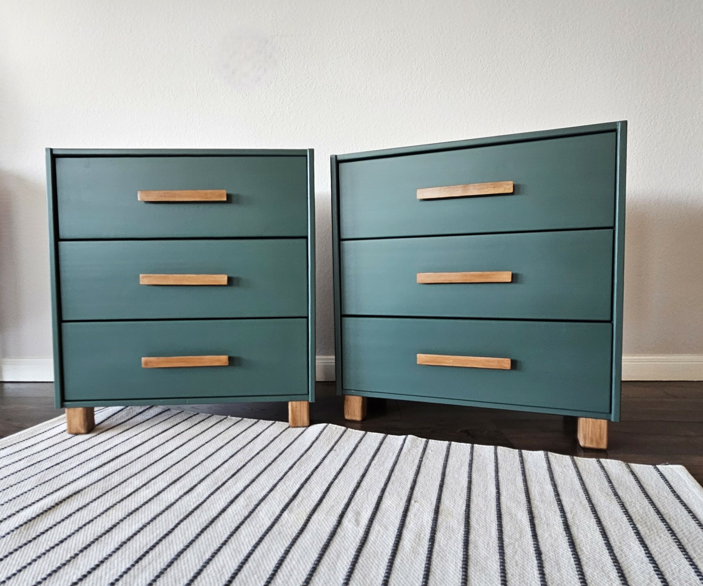 Two teal-colored dressers with wooden handles and legs, placed side by side on a striped rug against a white wall.