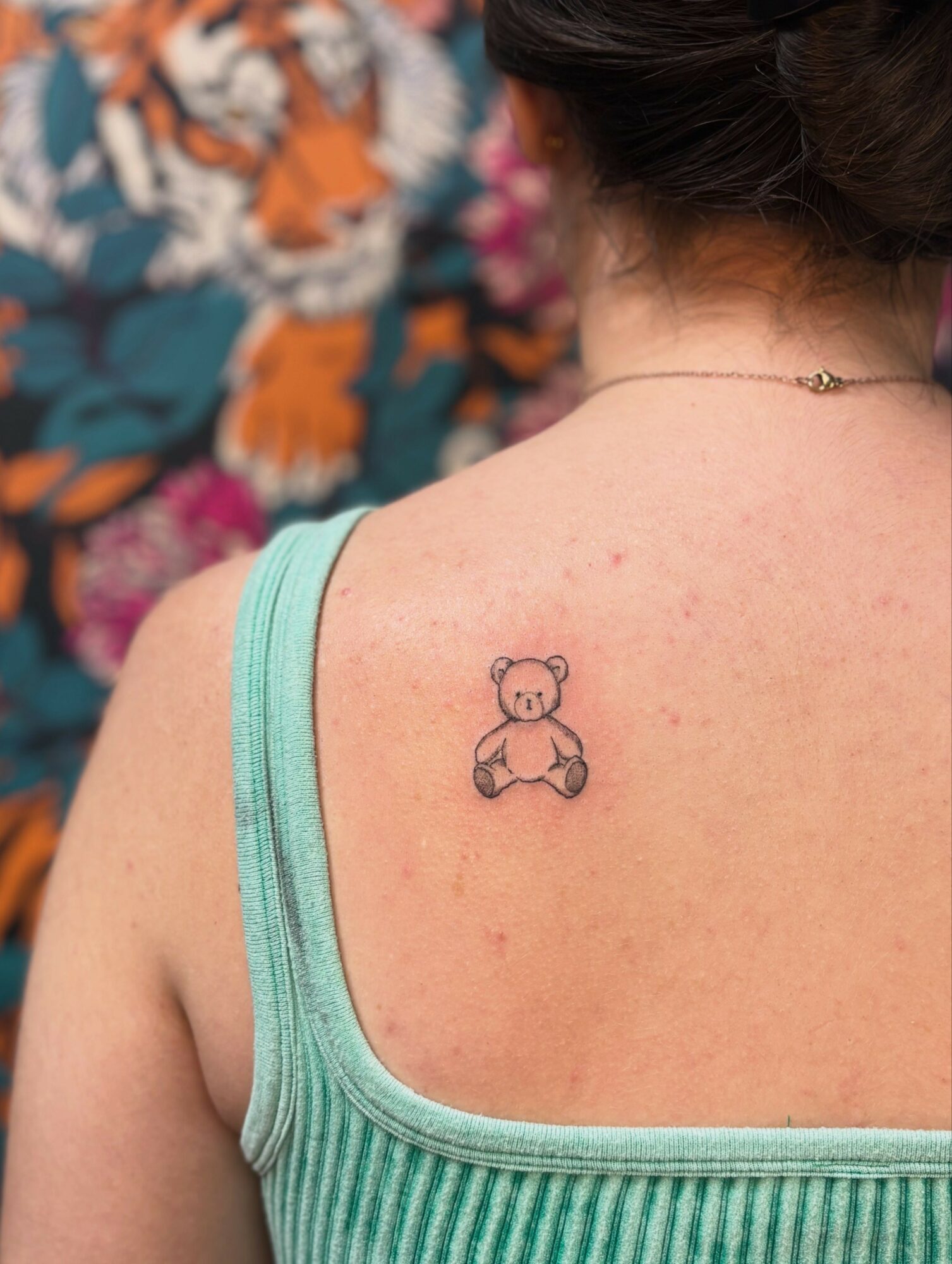 Person with a small teddy bear tattoo on shoulder, wearing a green tank top, with a floral background.