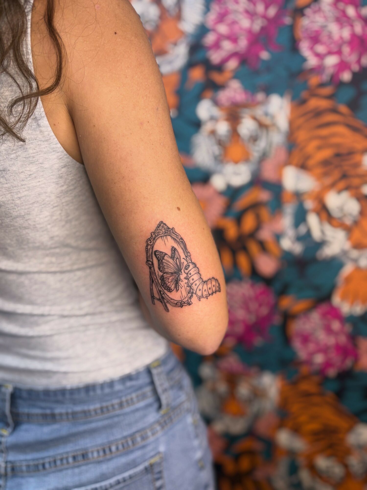 Person with a tattoo of a tiger on their upper arm, wearing a gray tank top and blue jeans, with a colorful floral background.