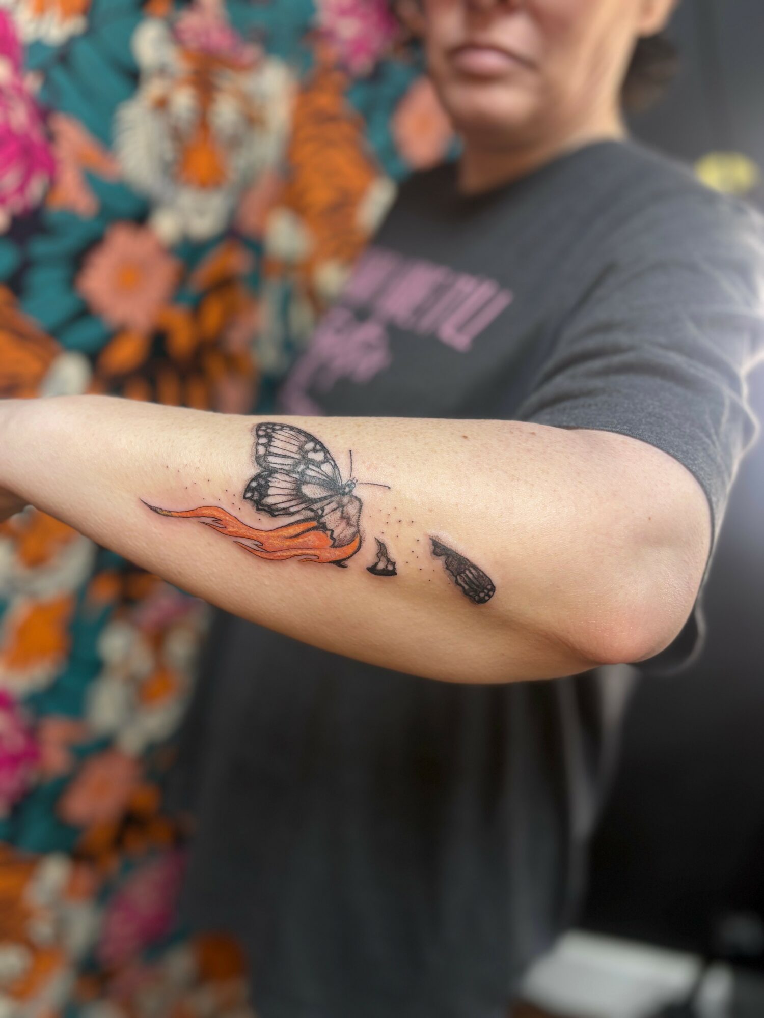 Person with a butterfly tattoo on their arm, with a colorful floral background, wearing a dark T-shirt.