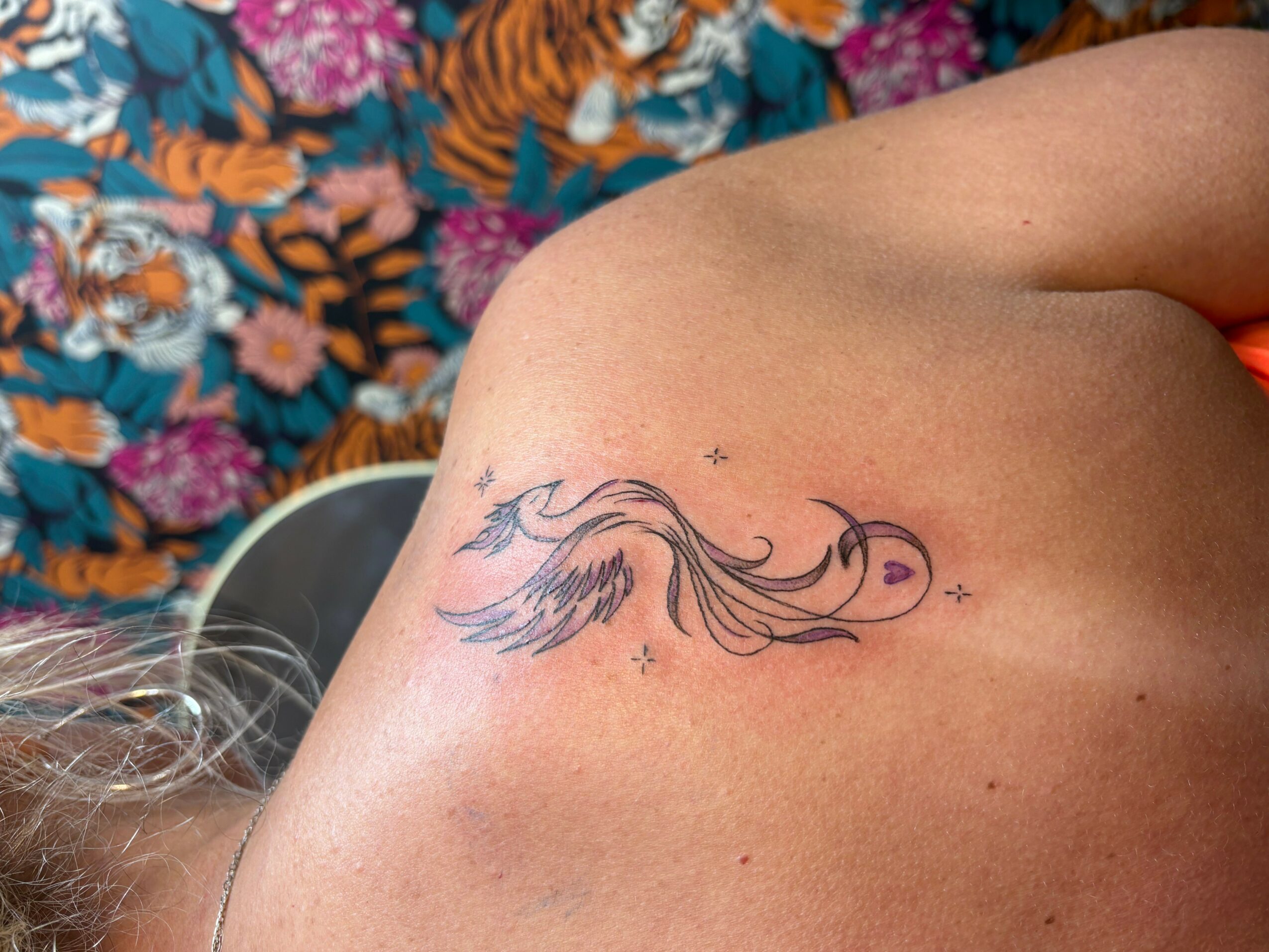 Tattoo of a stylized bird with flowing tail feathers on person's shoulder blade, with small stars around it.