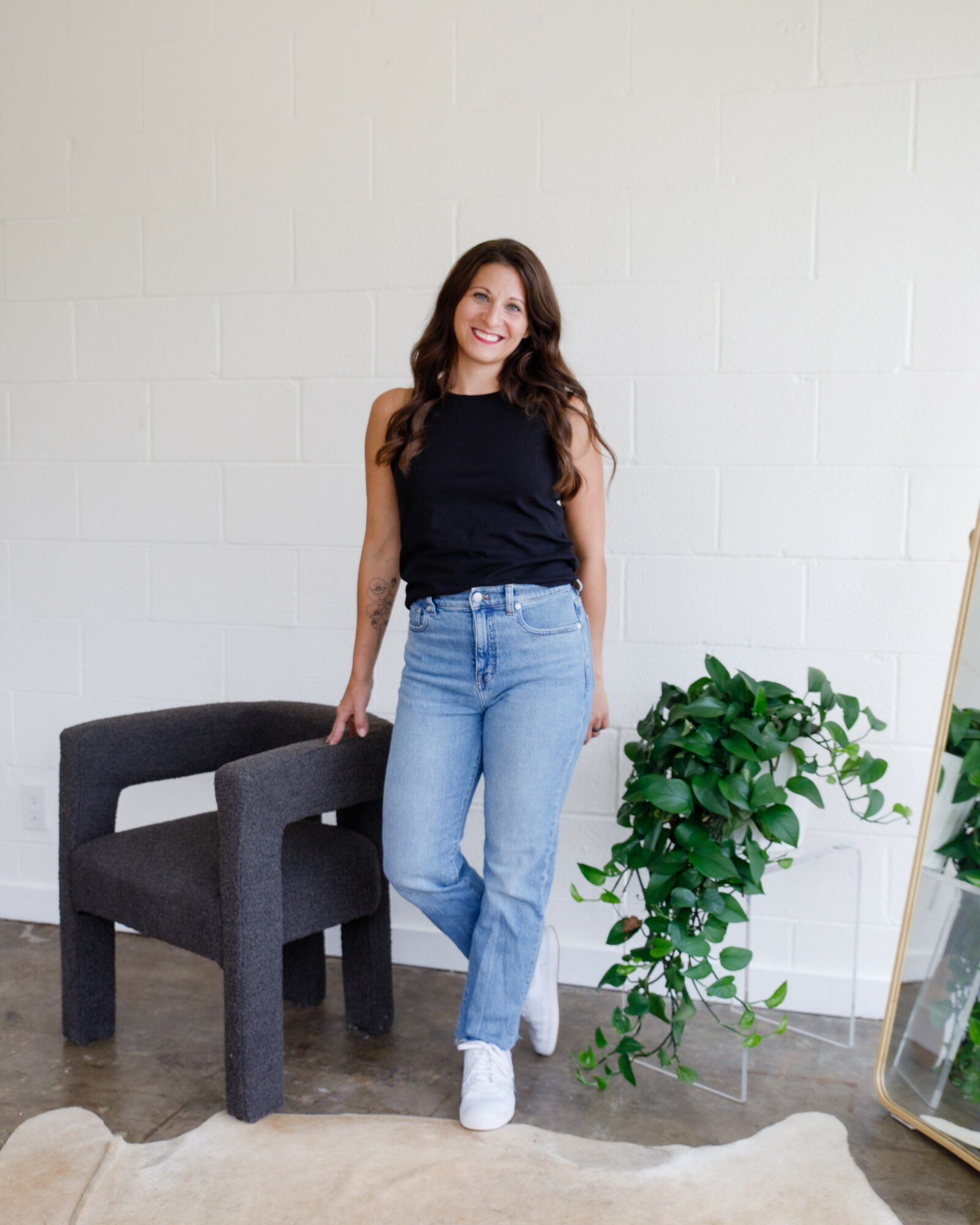 Woman with long hair in a black top and jeans standing indoors next to a black chair and a green plant.