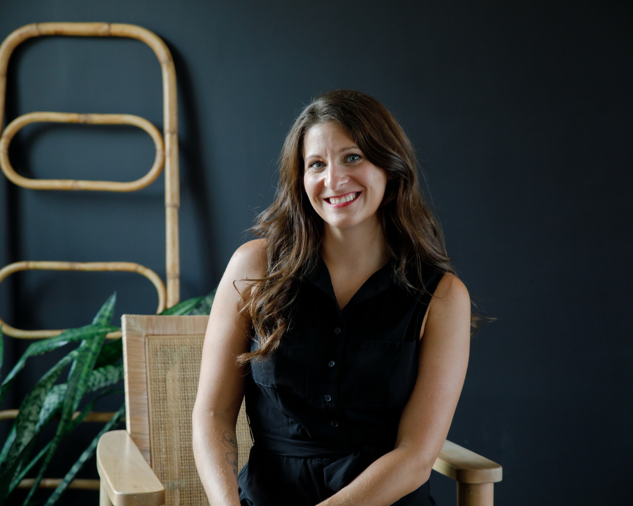 Woman with long brown hair smiling, sitting on a chair in front of a dark background with a plant and wooden structure