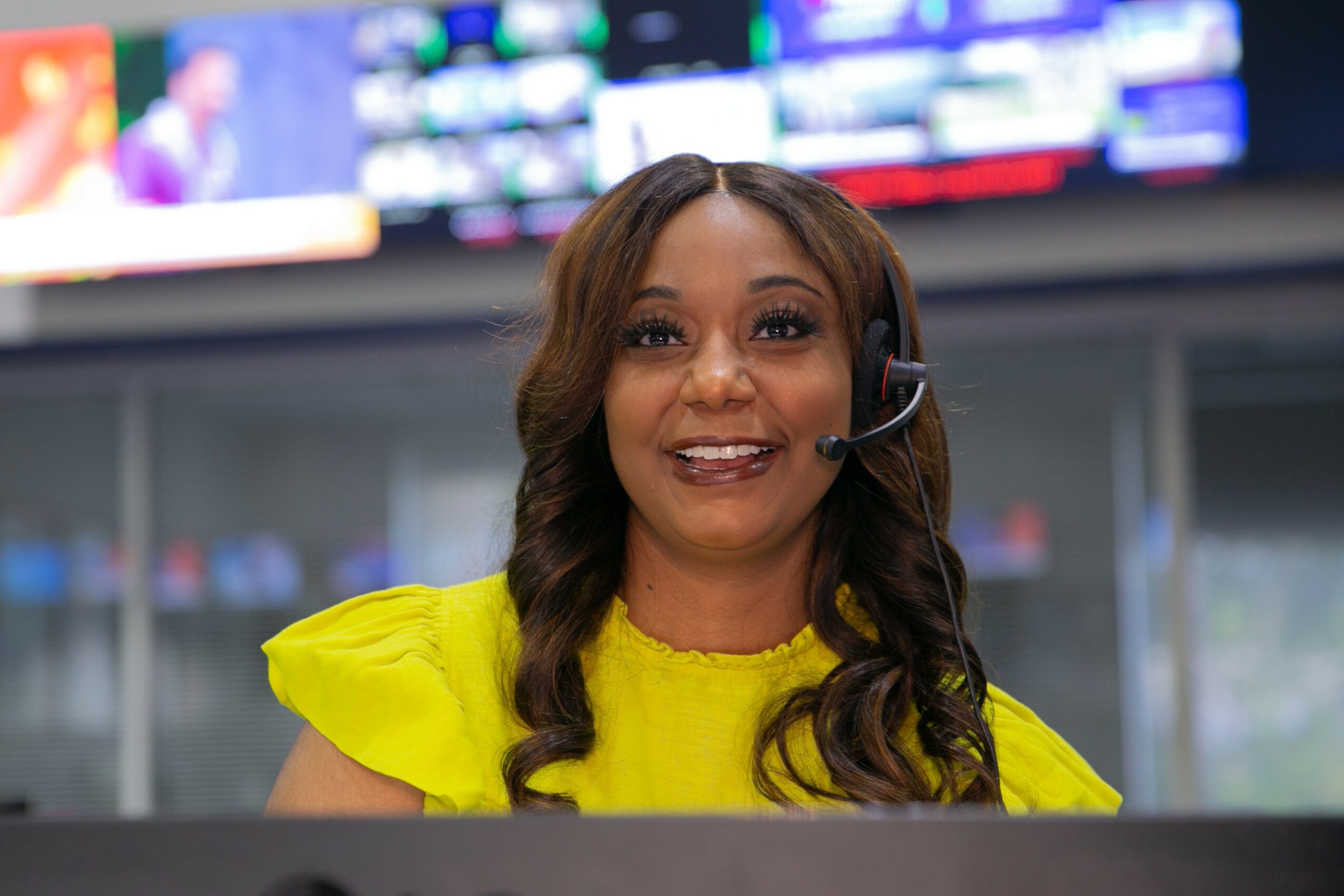 Woman with long wavy hair wearing a yellow top and headset, smiling, in a control room with screens behind her.