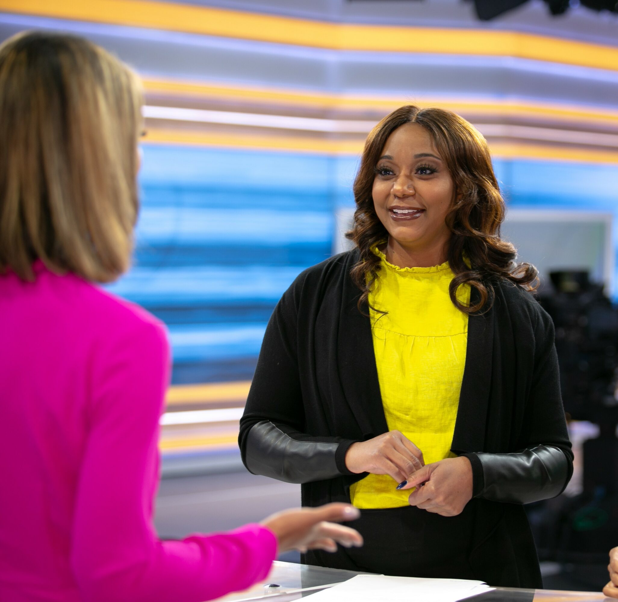 Two women talking in a brightly lit studio, one smiling, the other with back to camera, colorful background.