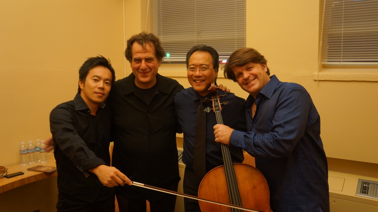 Four men standing together indoors, one holding a cello, smiling at the camera.