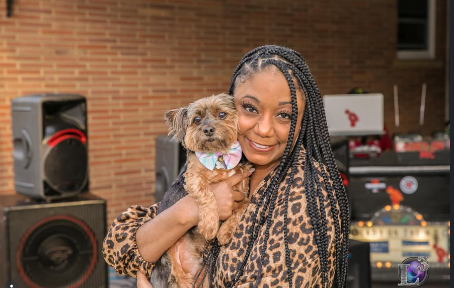 Woman with braided hair holding a small dog outdoors, with speakers and a brick wall in background.
