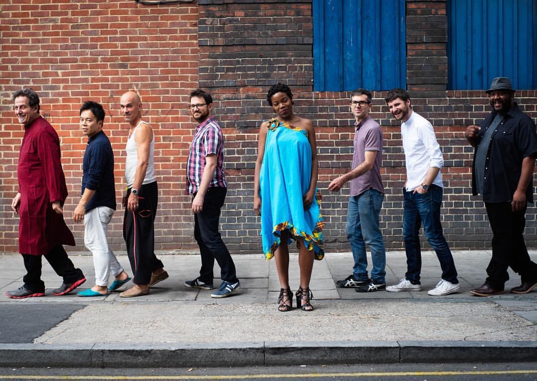 Group of nine people walking along a sidewalk in front of a brick wall with blue doors, one woman in a bright blue dress.