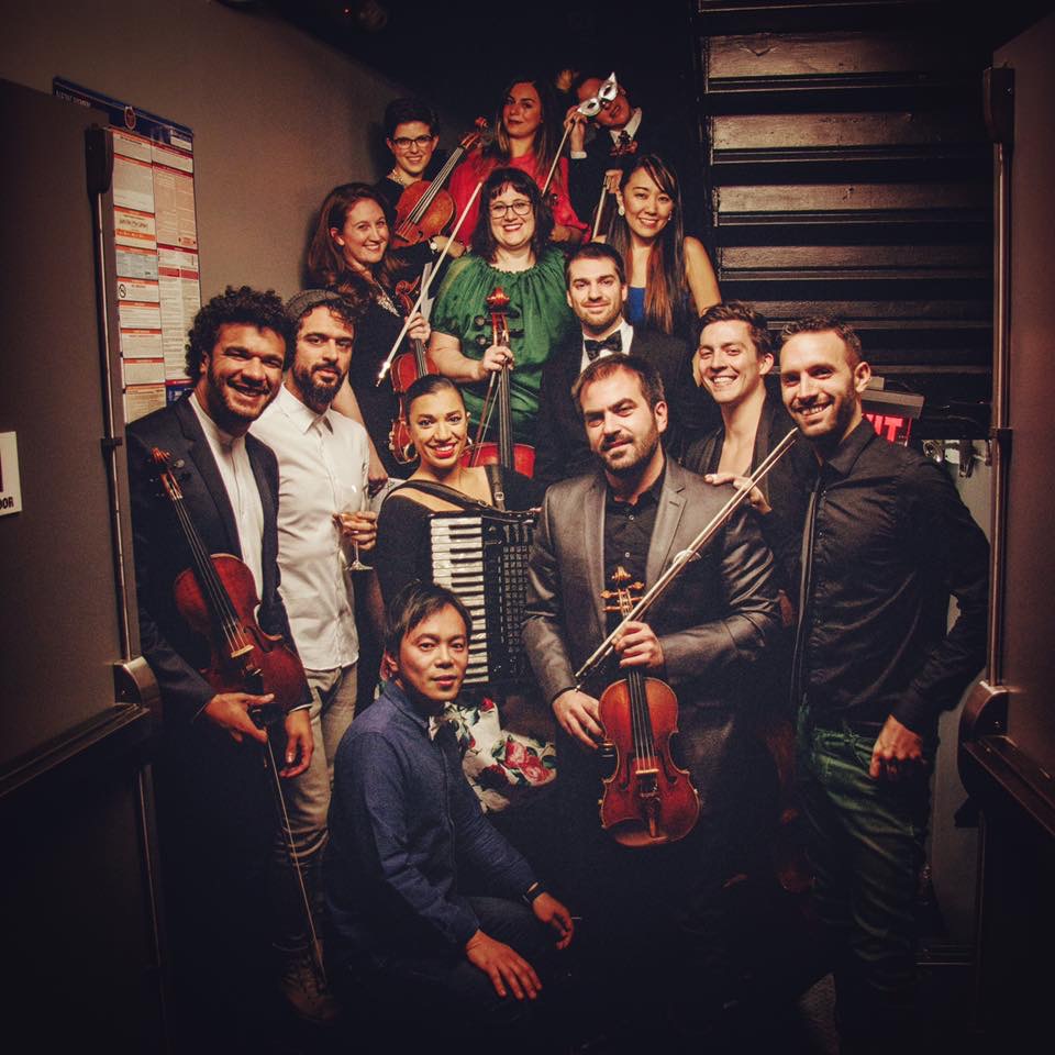 Group of musicians and performers posing indoors, some holding instruments, smiling, with stairs in the background.