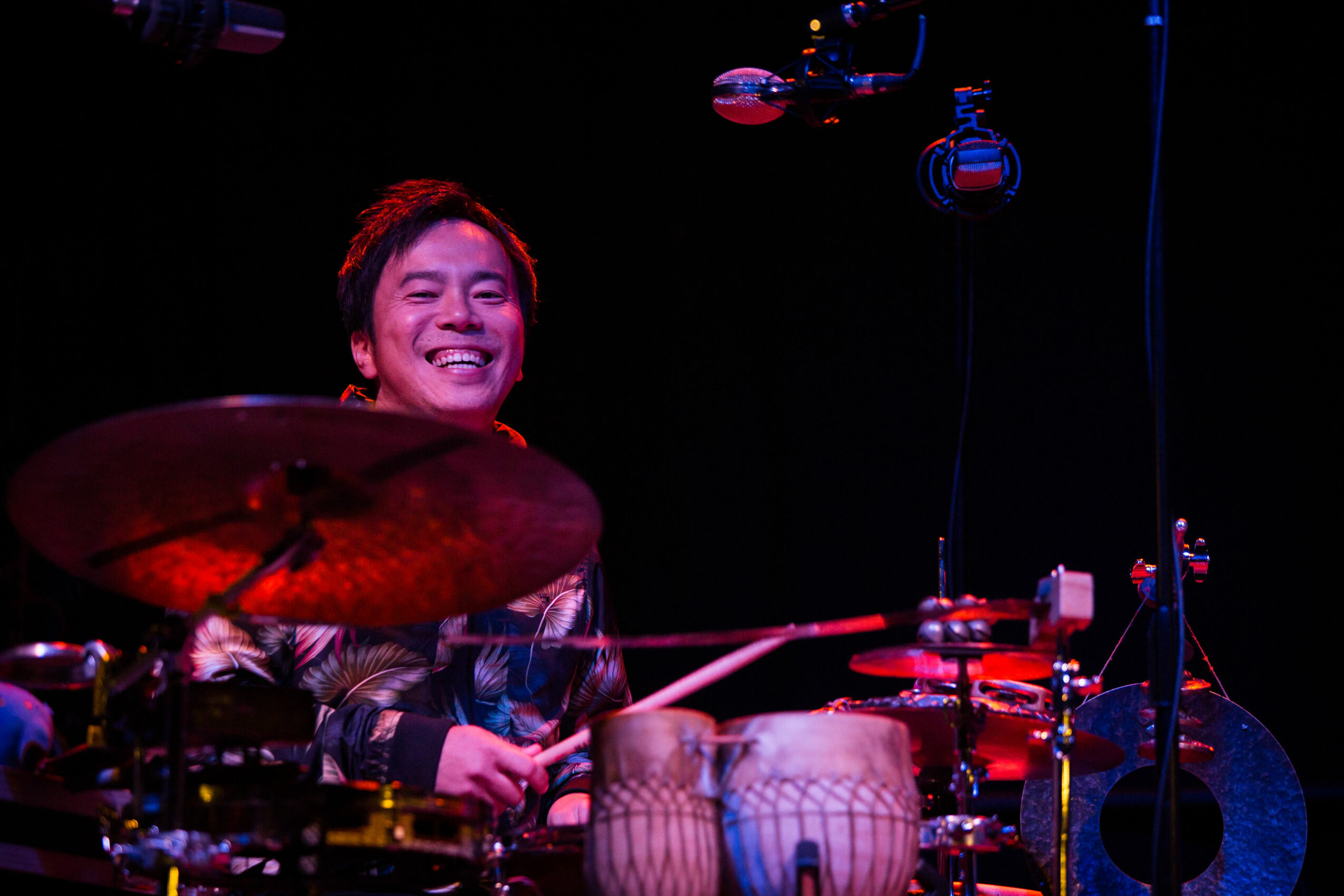 Person smiling while playing drums on stage with dark background.