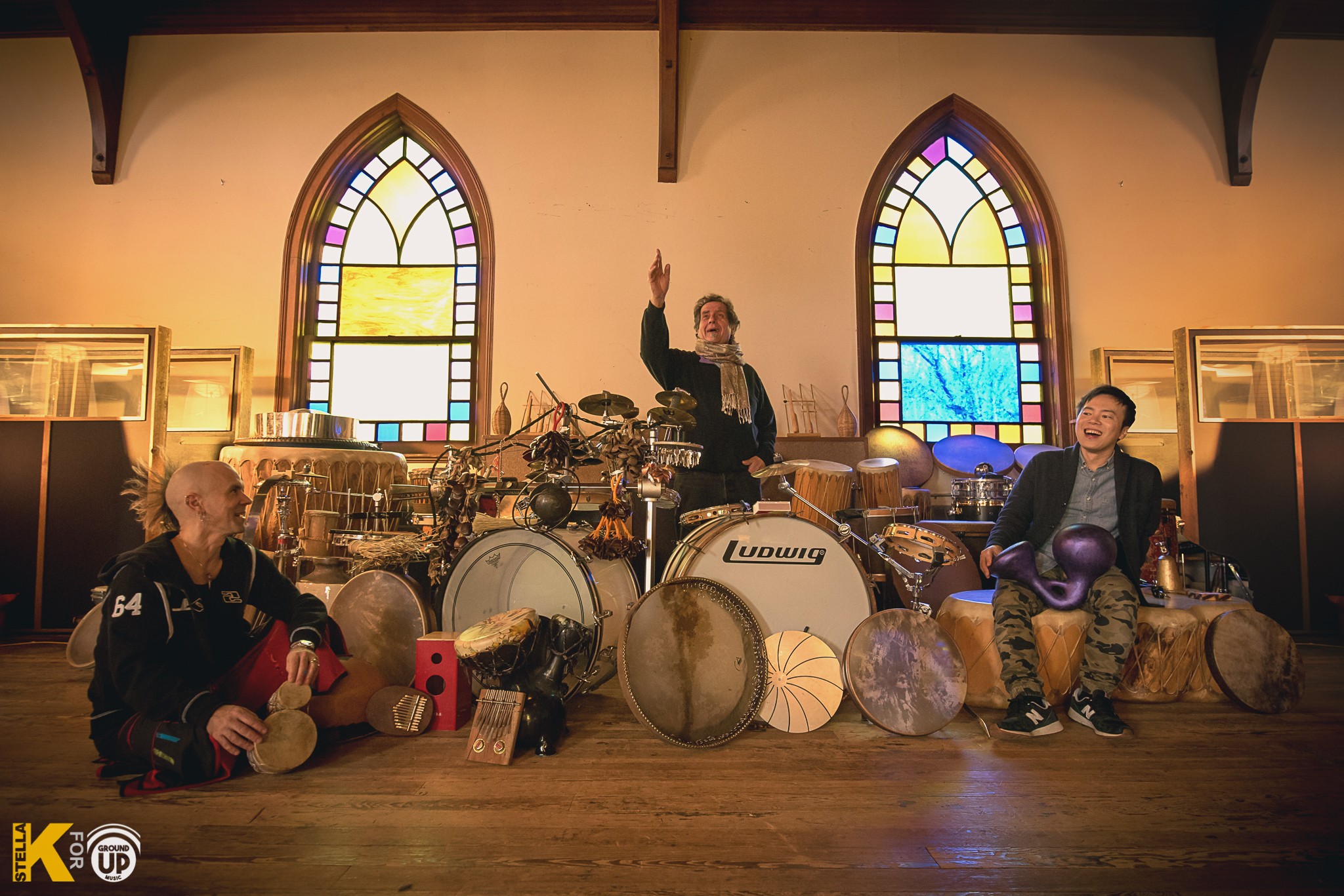 Three children sitting on the floor in front of a drum set inside a room with stained glass windows, one child raising a hand.