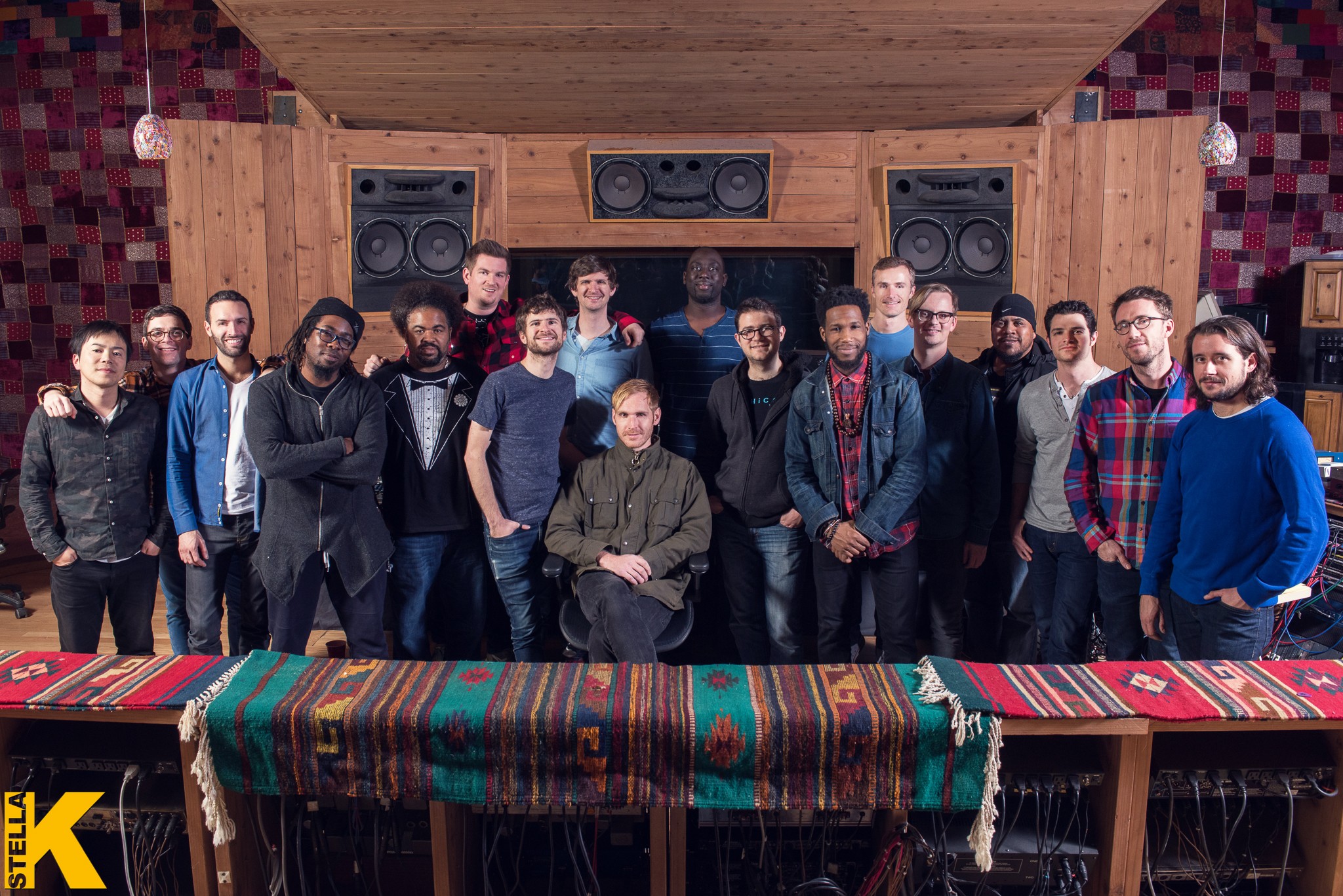 Group of 16 people in a recording studio with speakers, wooden walls, and a colorful tablecloth, posing for a photo.