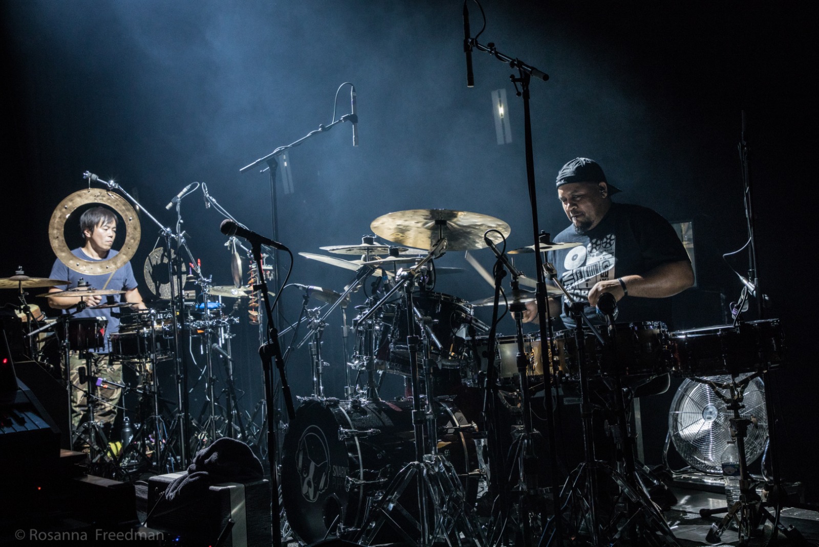 Two musicians perform on stage with drums and percussion instruments, illuminated by stage lights in a dark setting.