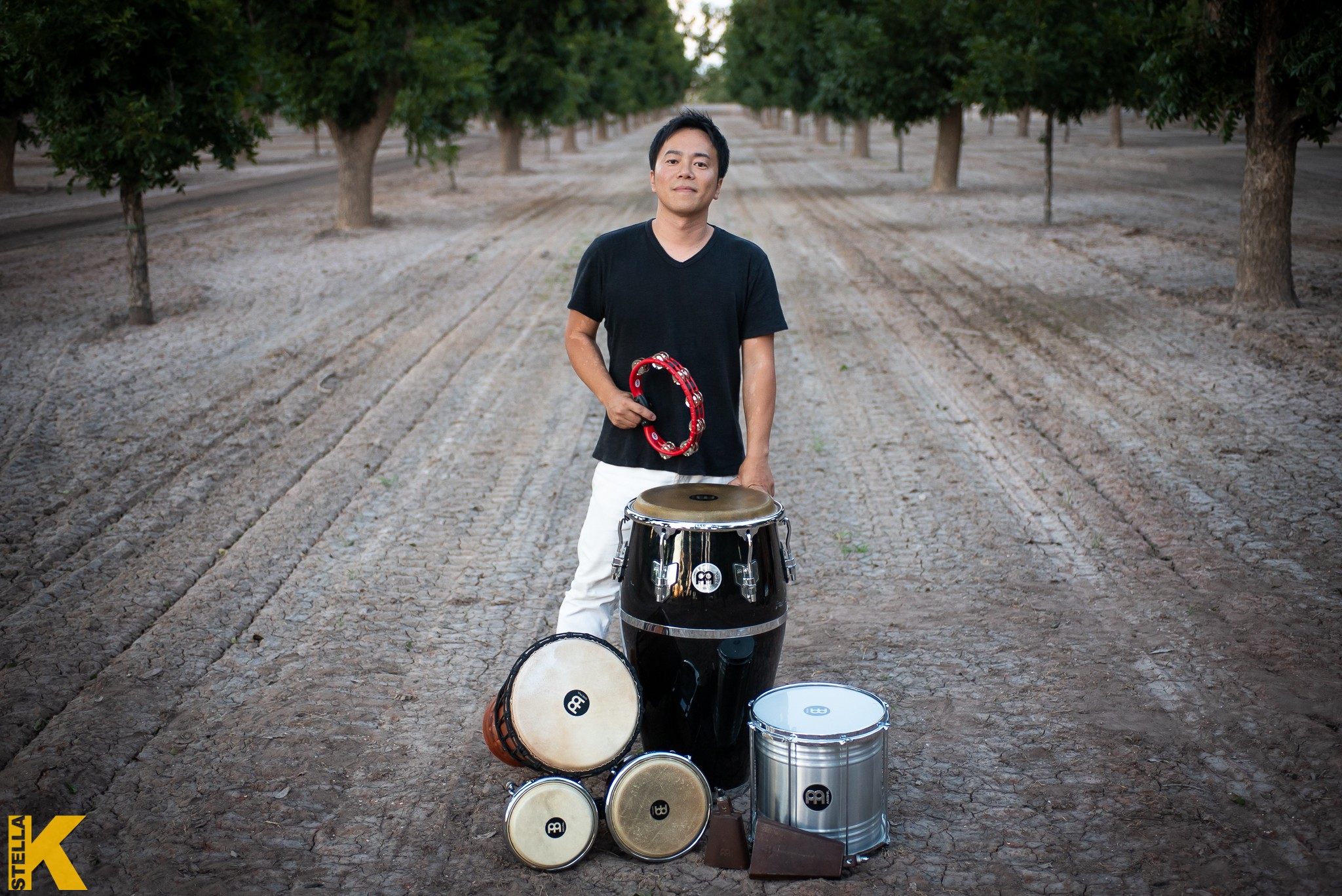 Person standing outdoors with drums, trees in background, holding drumsticks, on a dirt path.