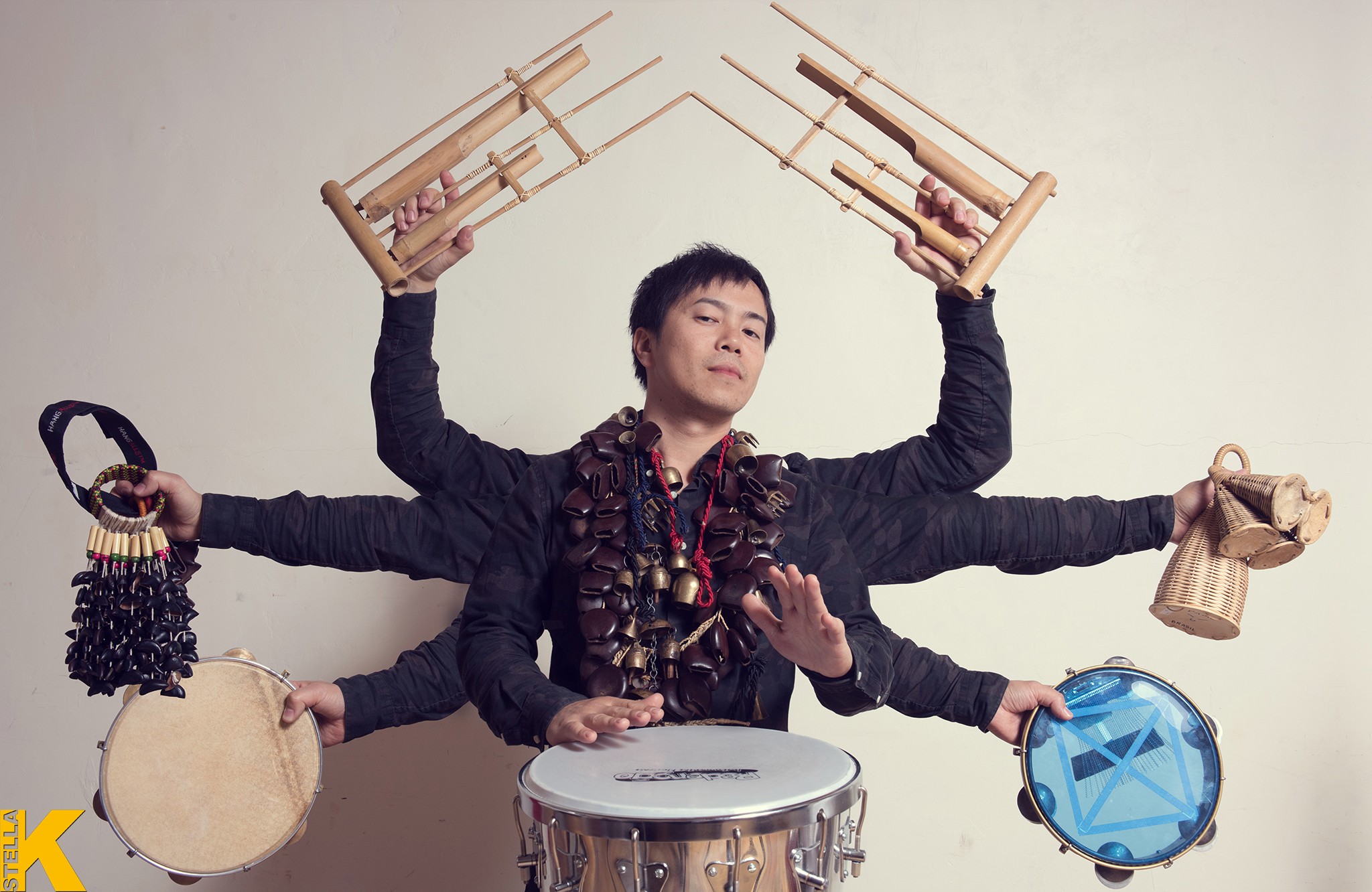 Person with multiple arms holding musical instruments and objects, performing with a drum, against a plain background.
