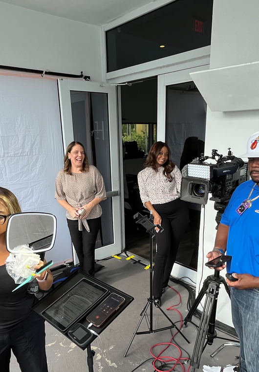 Two women stand near a camera and equipment in a room with large windows and a white backdrop.