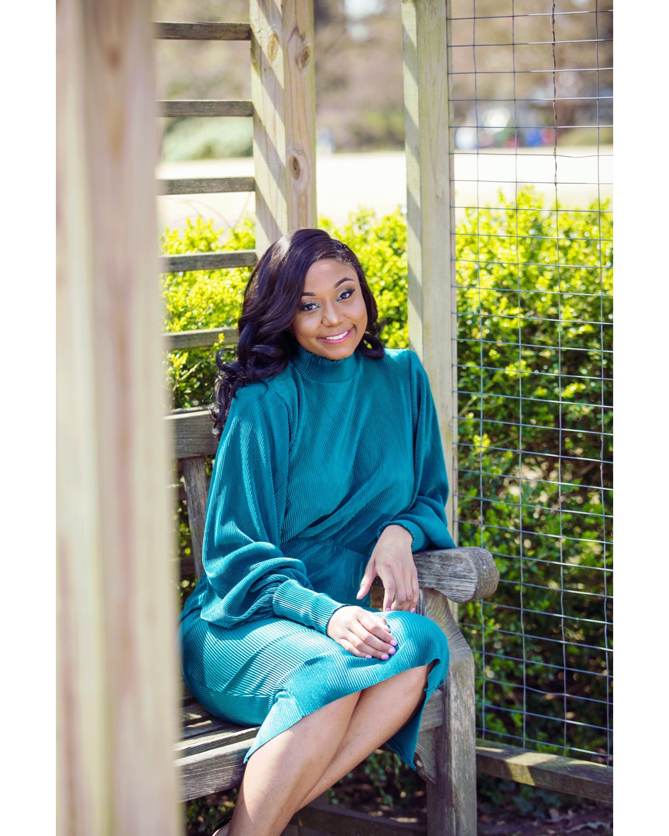 Woman sitting on a wooden bench outdoors, smiling, wearing a teal dress, surrounded by greenery and wooden structures.