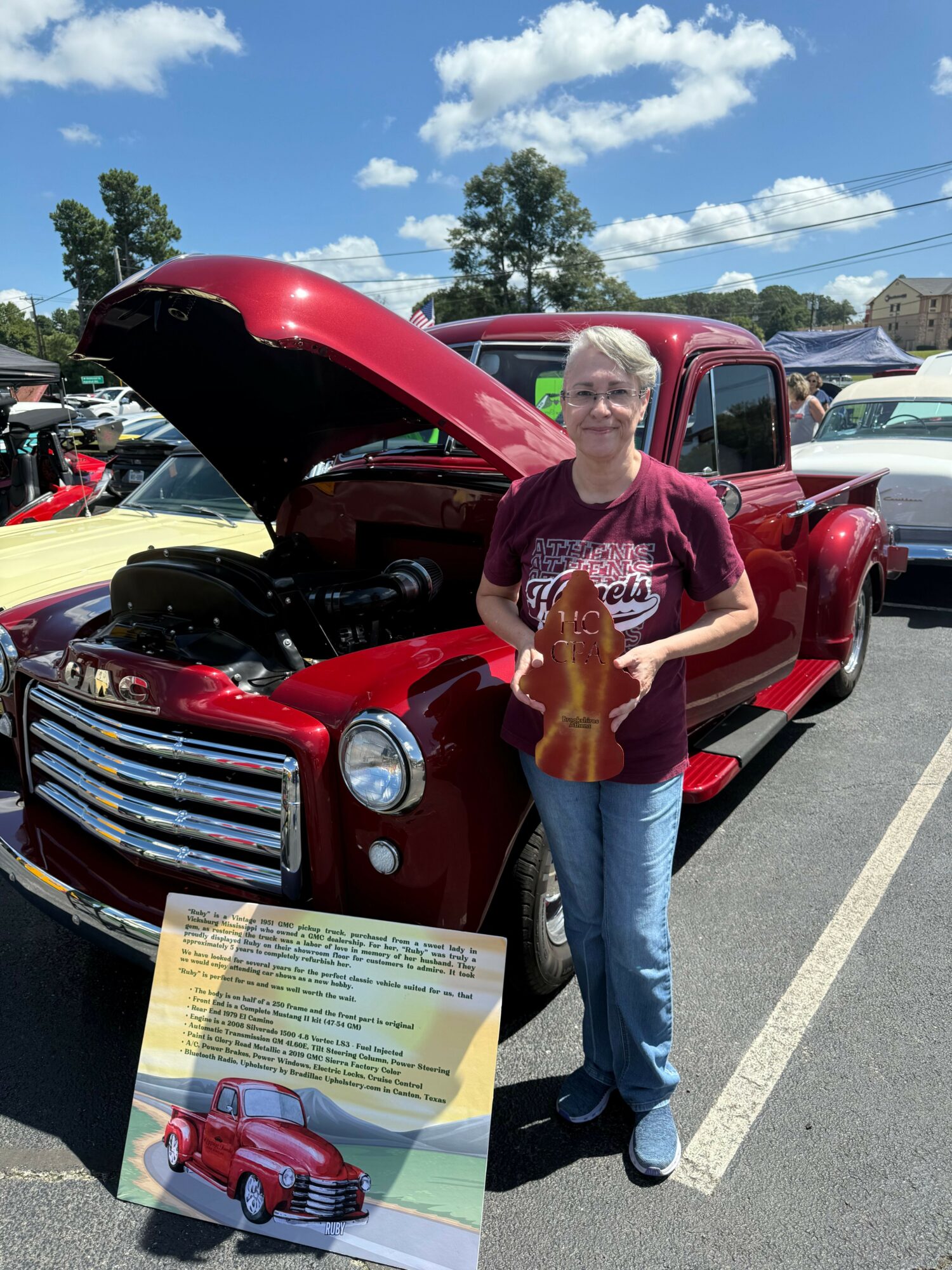 Person with long hair holding a bag, standing next to a red vintage car at an outdoor event, with a brochure on the ground.