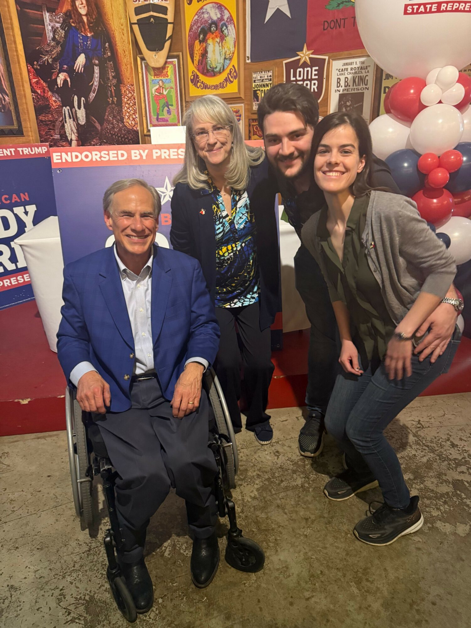 Three people posing indoors, one in a wheelchair, with colorful balloons and posters in the background.