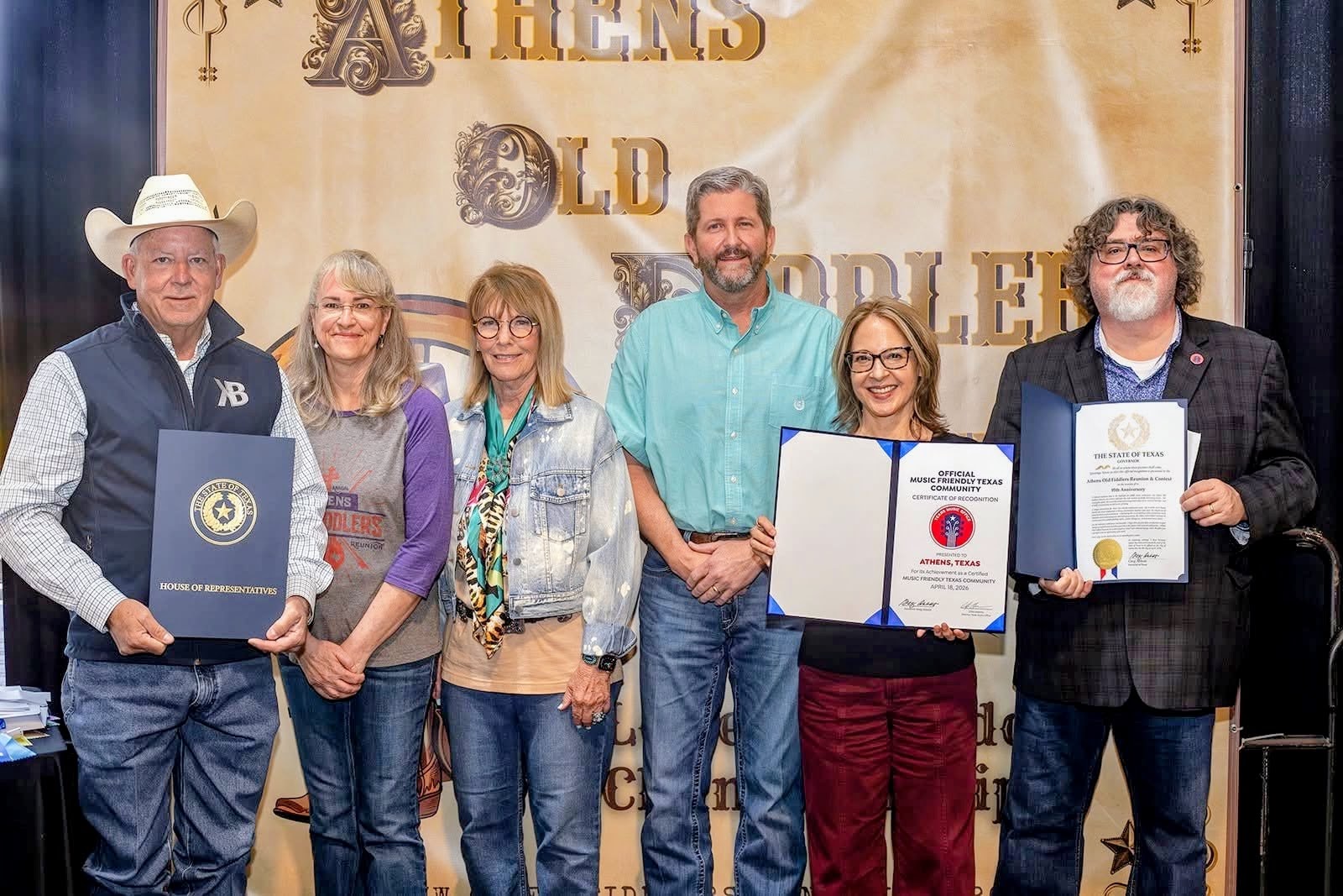 Group of six people standing in front of a banner, holding awards and certificates, smiling at the camera.