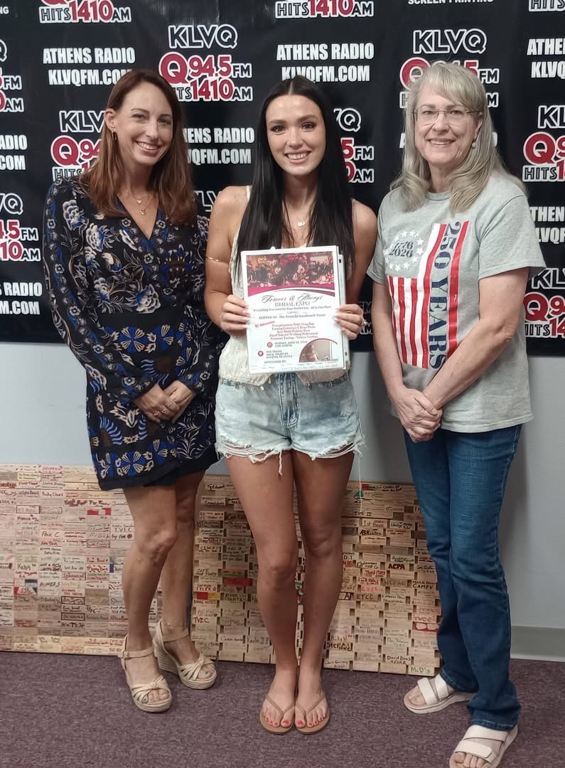 Three women standing in front of a radio station backdrop, one holding a certificate, smiling.