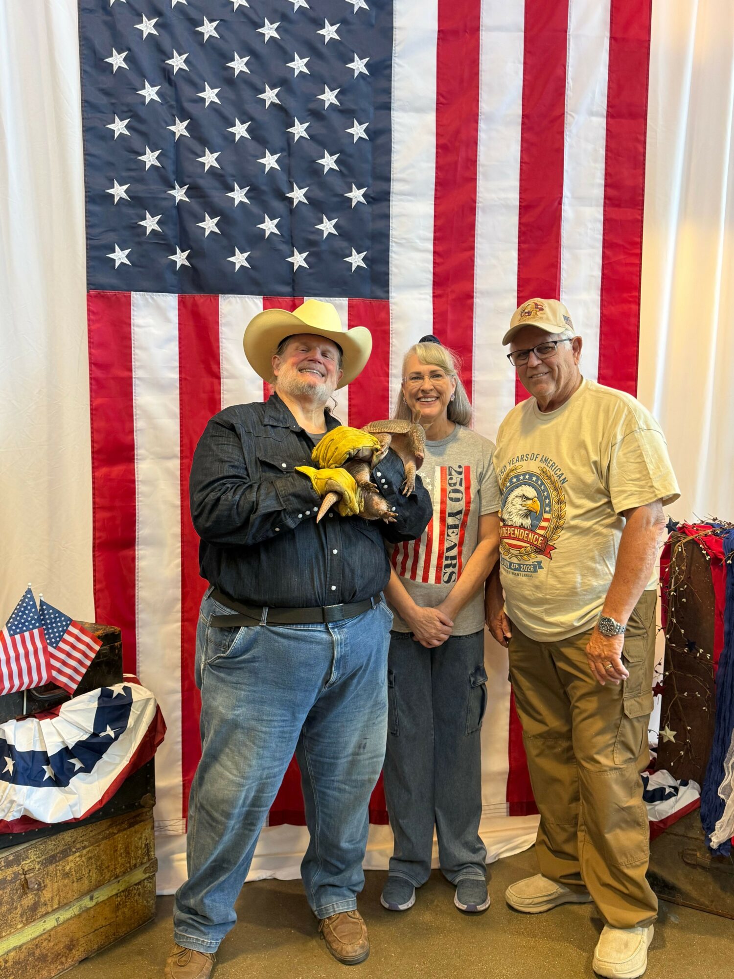 Three people standing in front of an American flag, with a child holding a small American flag, and a woman wearing a yellow hat and gloves. A patriotic display with a flag backdrop and a patriotic-themed hat on a table.