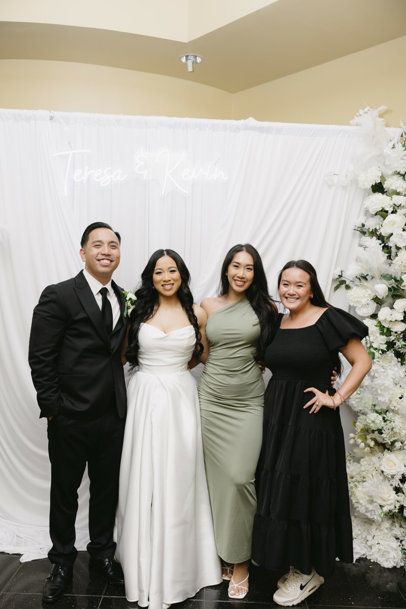 Four people standing in front of a white floral backdrop, smiling, dressed in formal attire, with a neon sign above.