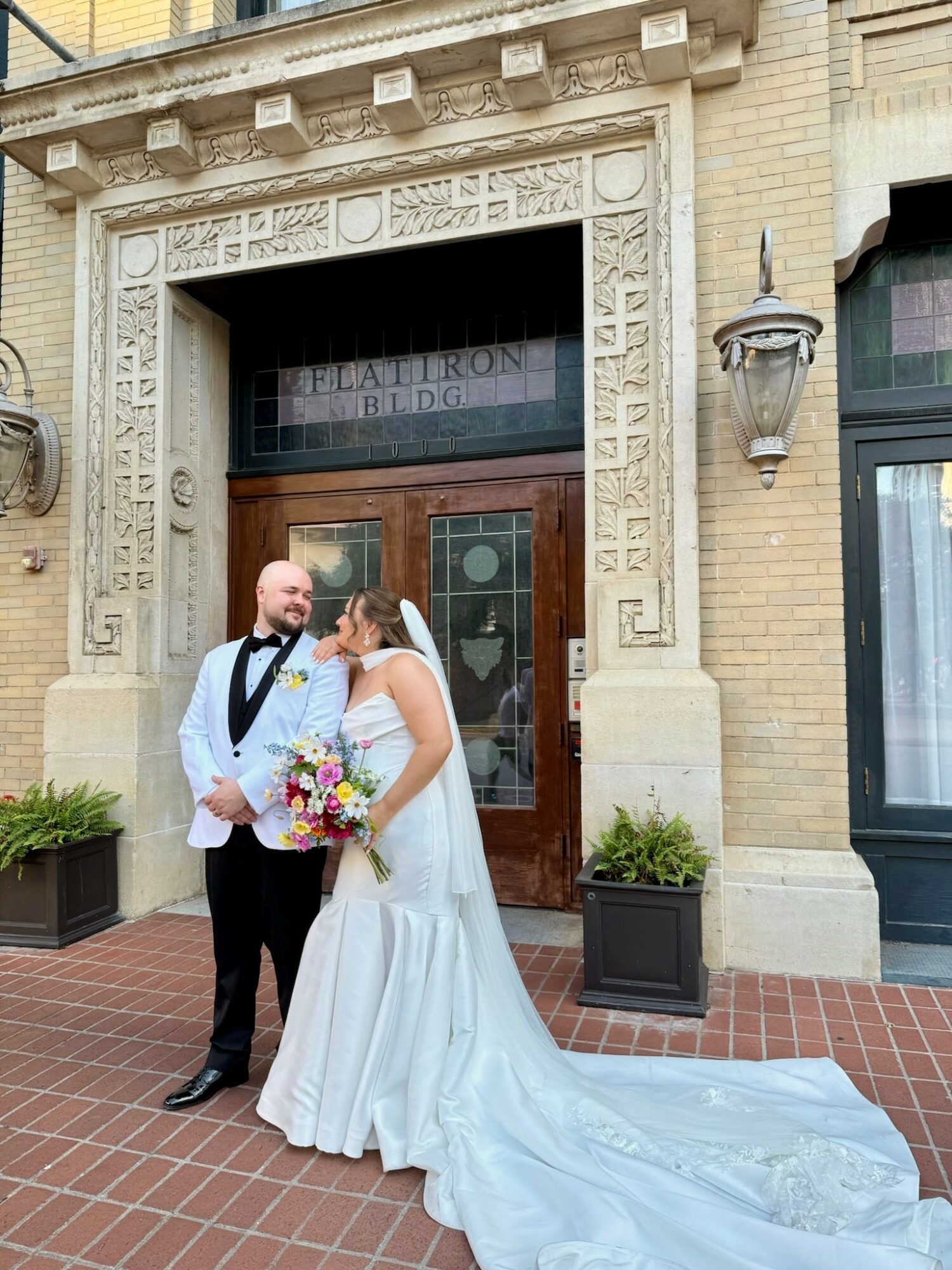 Bride and groom stand outside a building with ornate stonework, bride holding a bouquet, both smiling at each other.