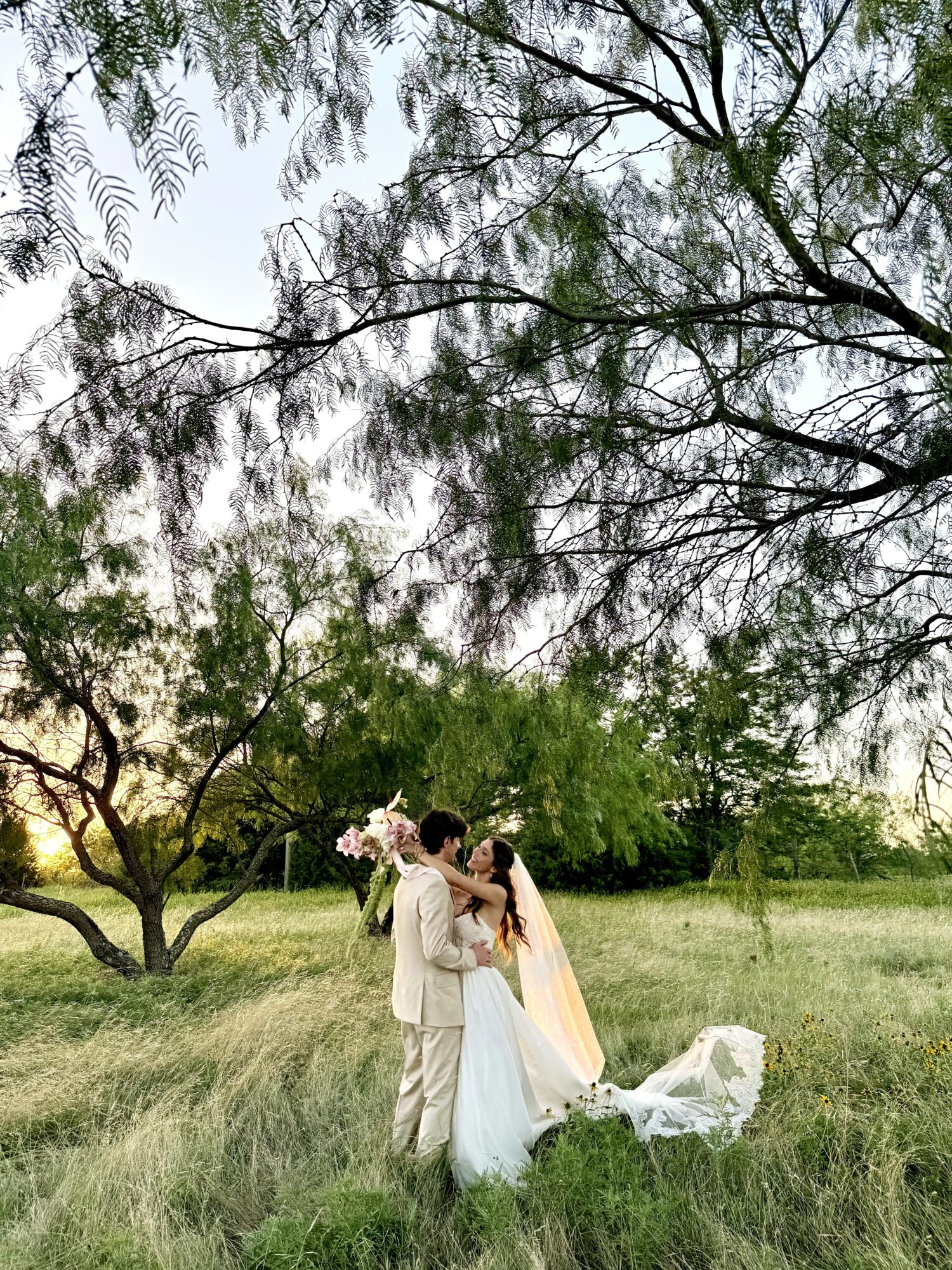 Two people in wedding attire standing in a grassy field with trees and sky in the background.
