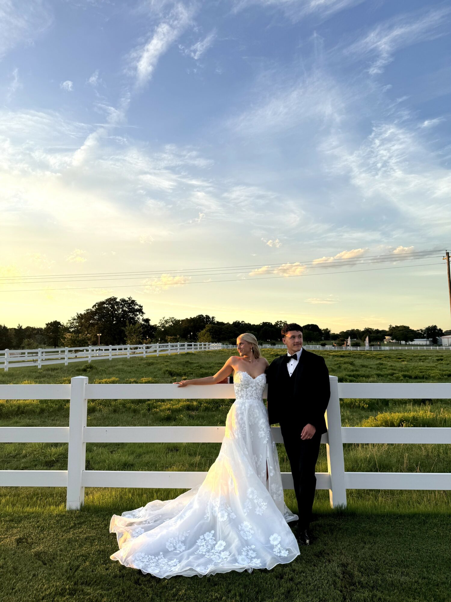 Bride and groom standing outdoors near a white fence, with a blue sky and clouds in the background.