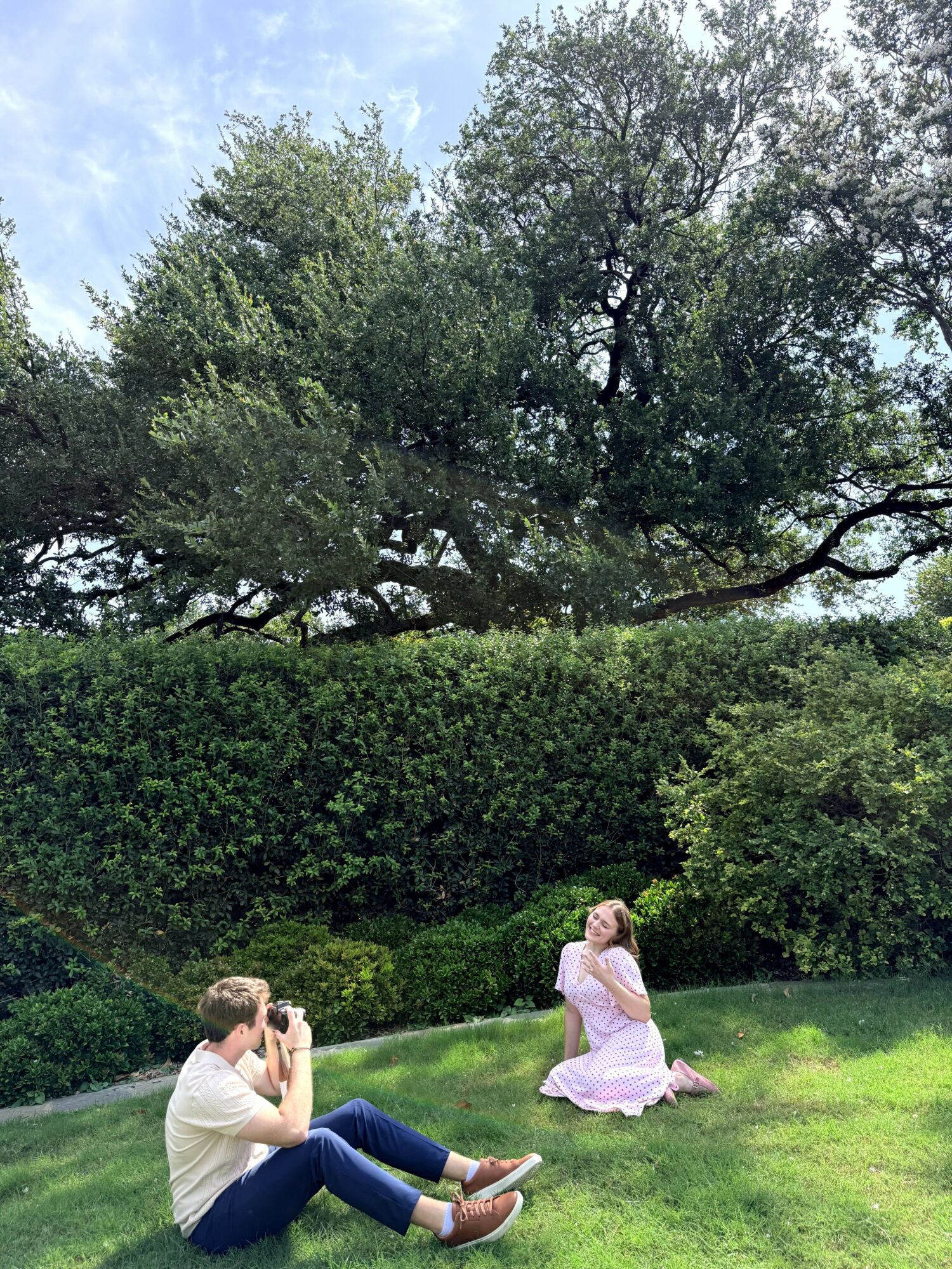 Person sitting on grass in a pink dress, looking up, with another person taking a photo nearby, surrounded by trees and bushes.