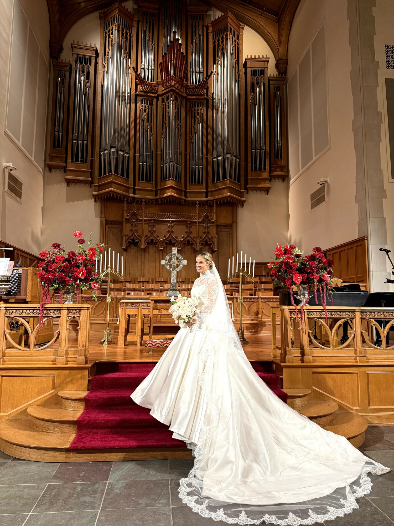 Bride in a white wedding gown holding a bouquet, standing on a red carpeted staircase inside a church with large pipe organ above.