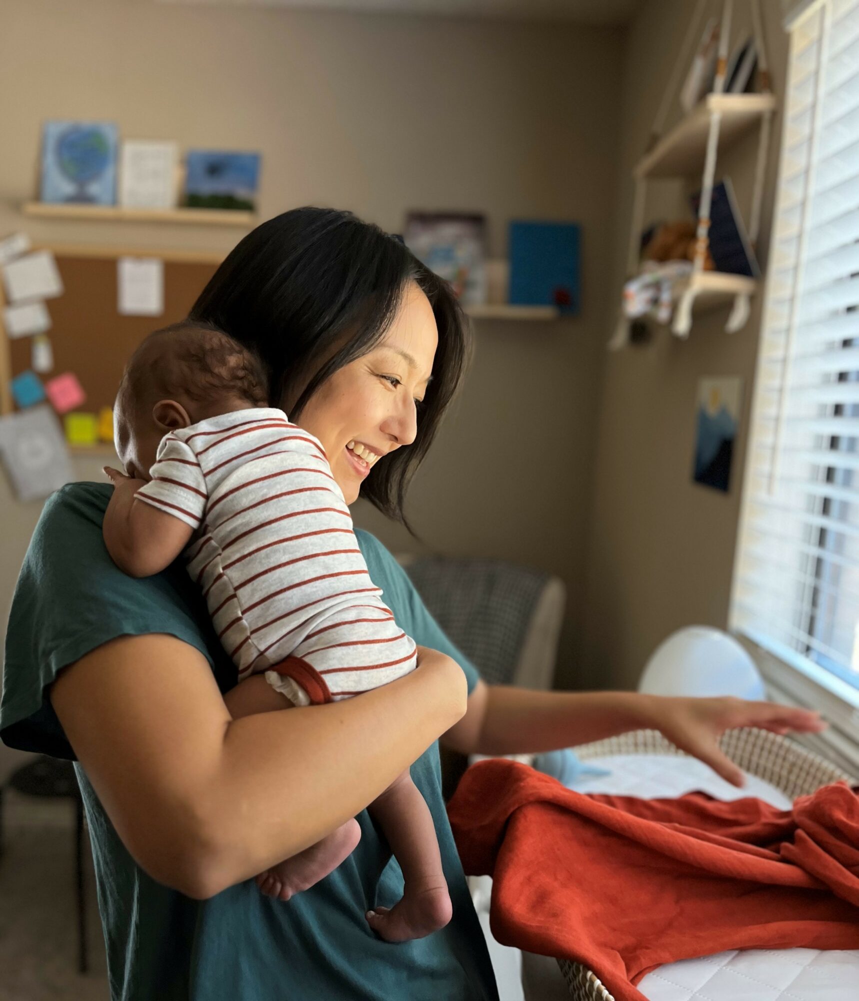 Woman holding a baby indoors, smiling, with shelves and window in background.