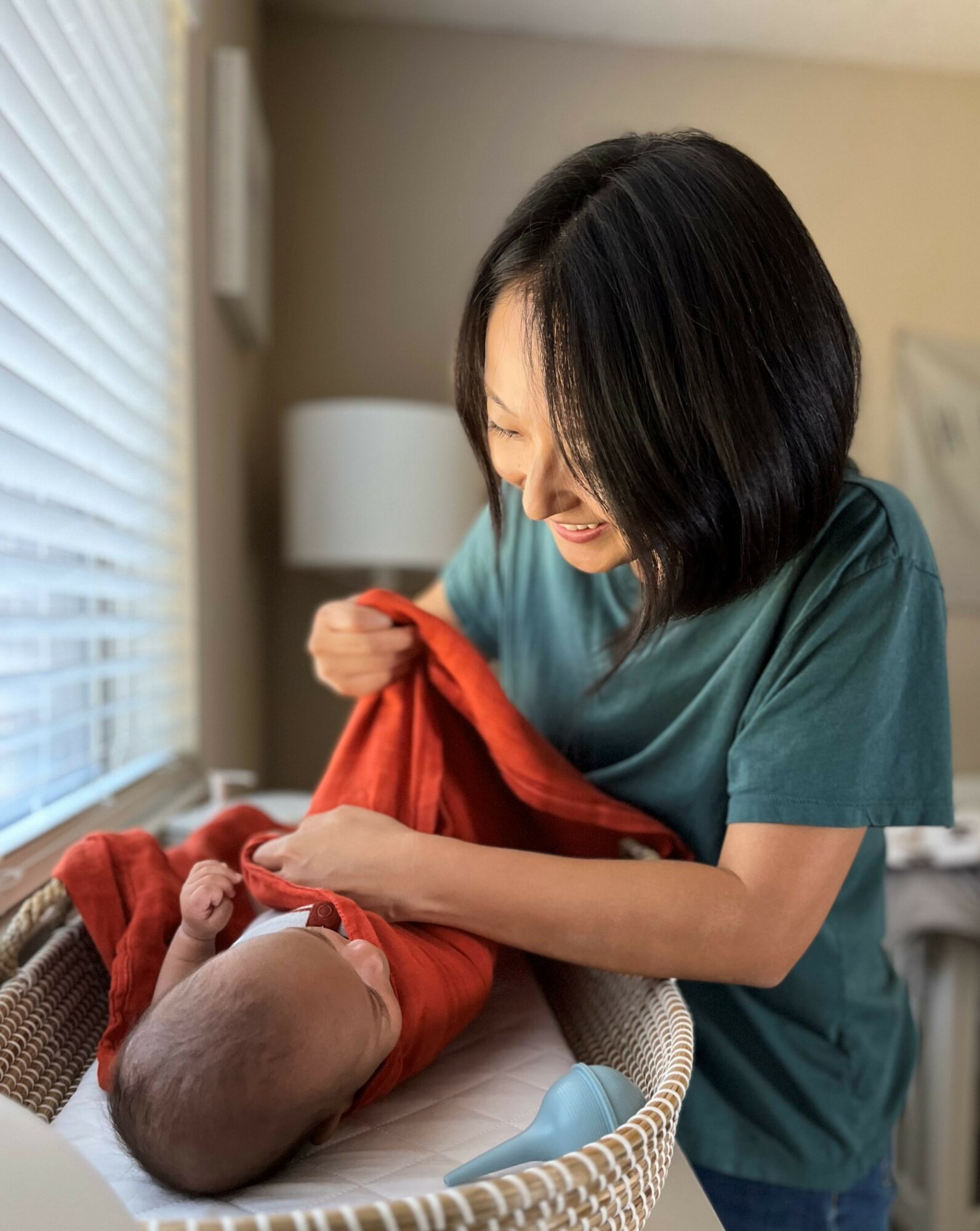 Woman smiling and holding a baby wrapped in a red blanket in a room with natural light.