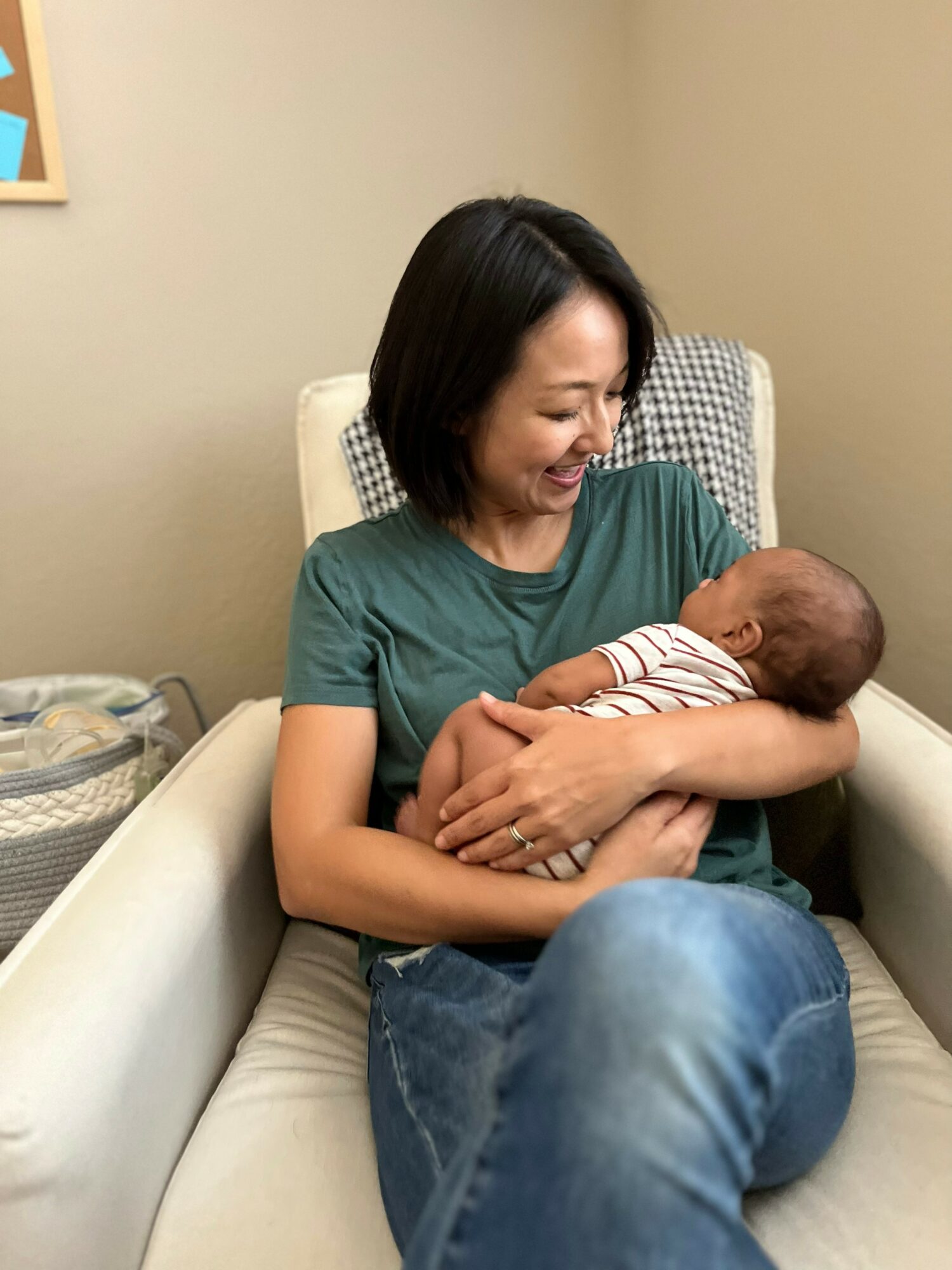 Woman holding a newborn baby, smiling, sitting on a chair in a room with beige walls.