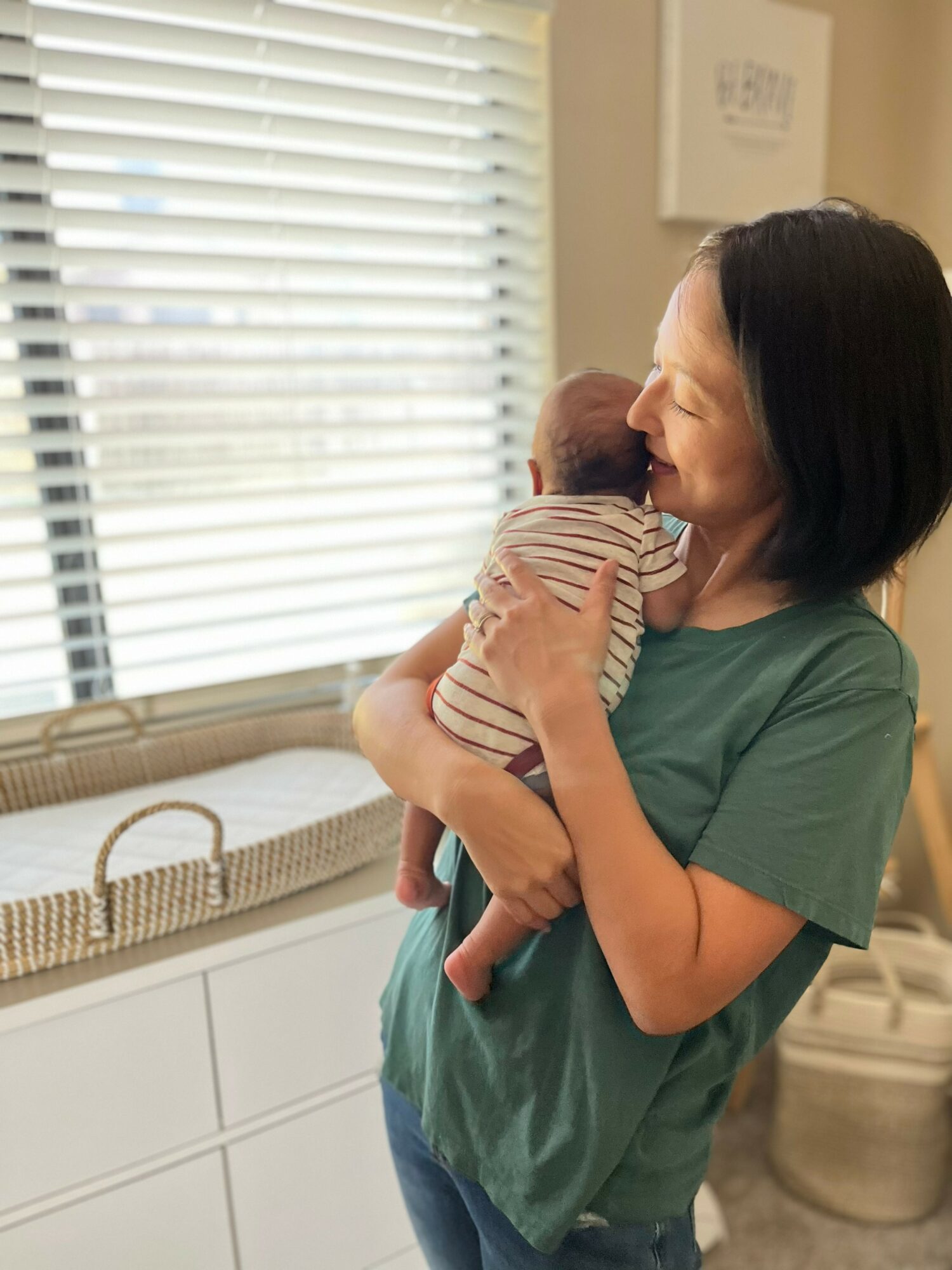 Woman holding a baby near a window with blinds, smiling and looking at the child.