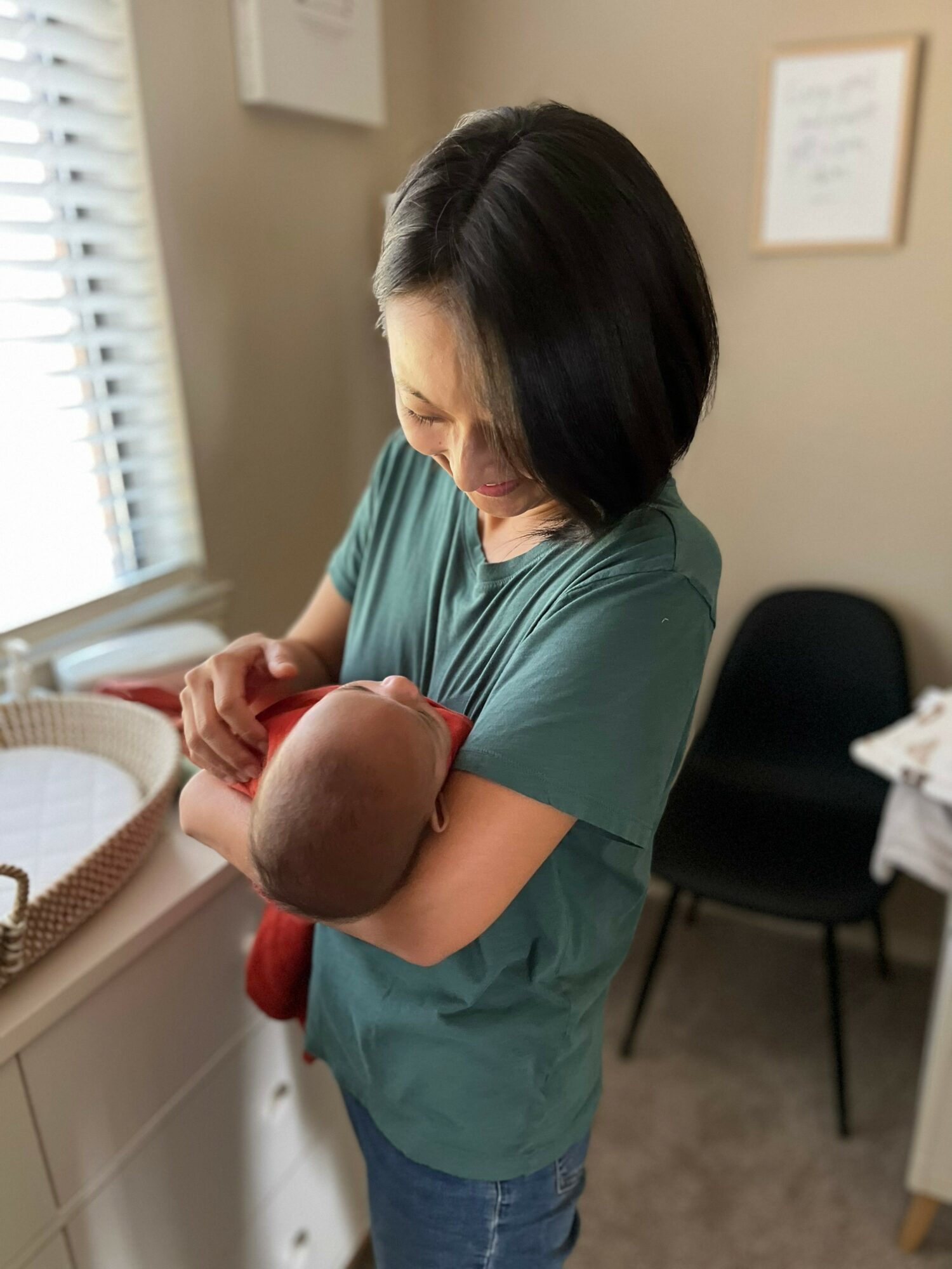 Woman holding a newborn baby near a window in a room with framed pictures on the wall.