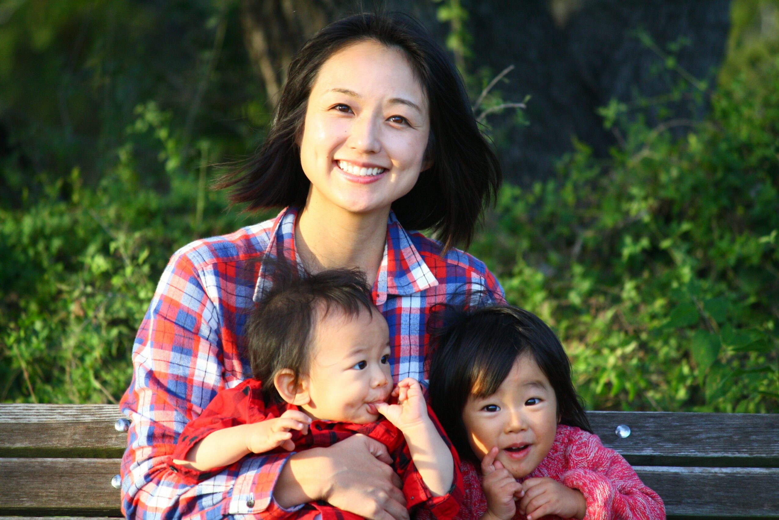 Smiling woman with two children sitting on a park bench, surrounded by green foliage.