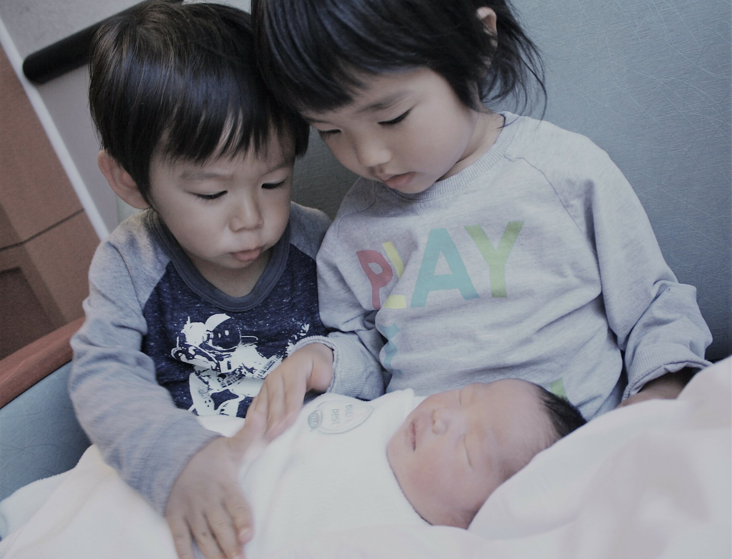 Two young children look at a newborn baby lying in a bed, with one child touching the baby's hand.