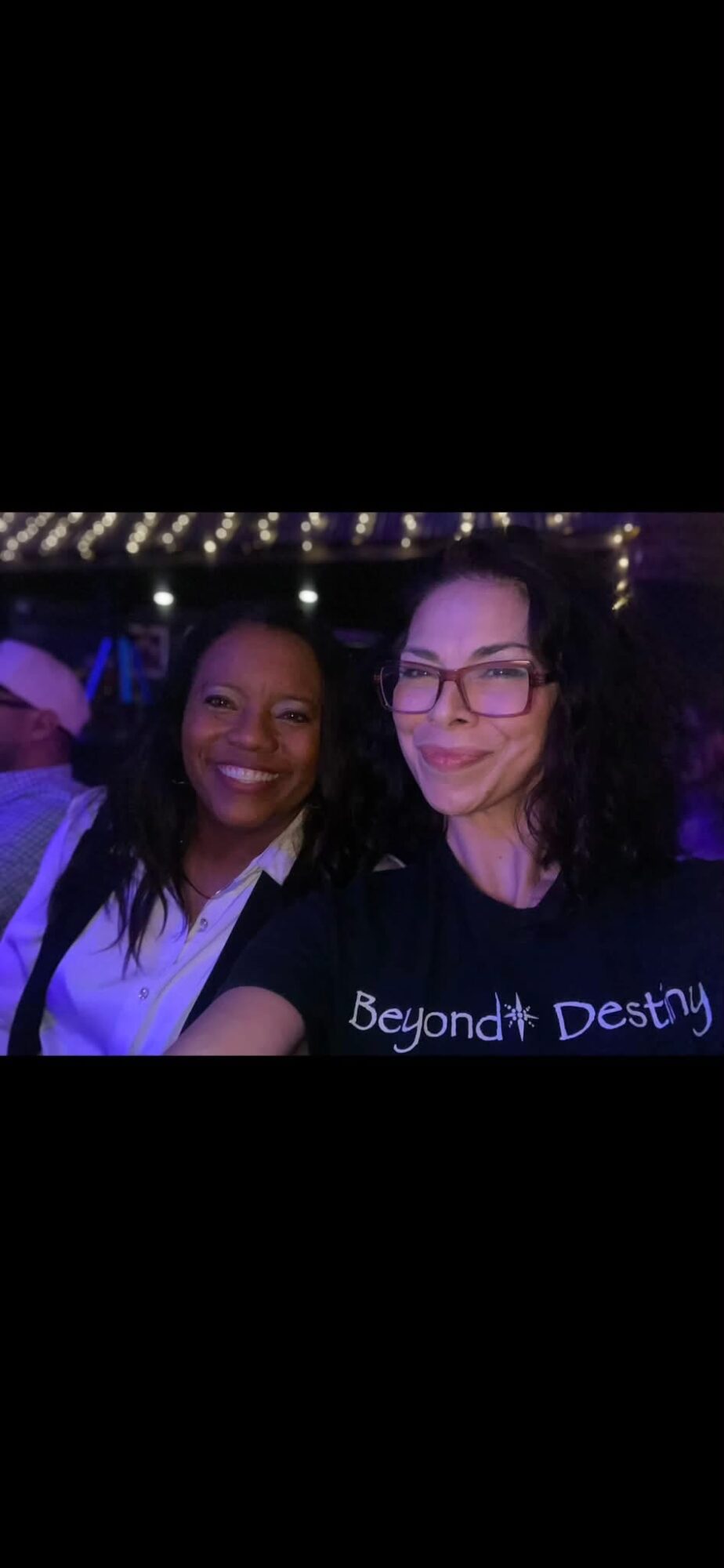 Two women smiling at a nighttime event with string lights overhead, one wearing glasses and a black shirt, the other in a white shirt.
