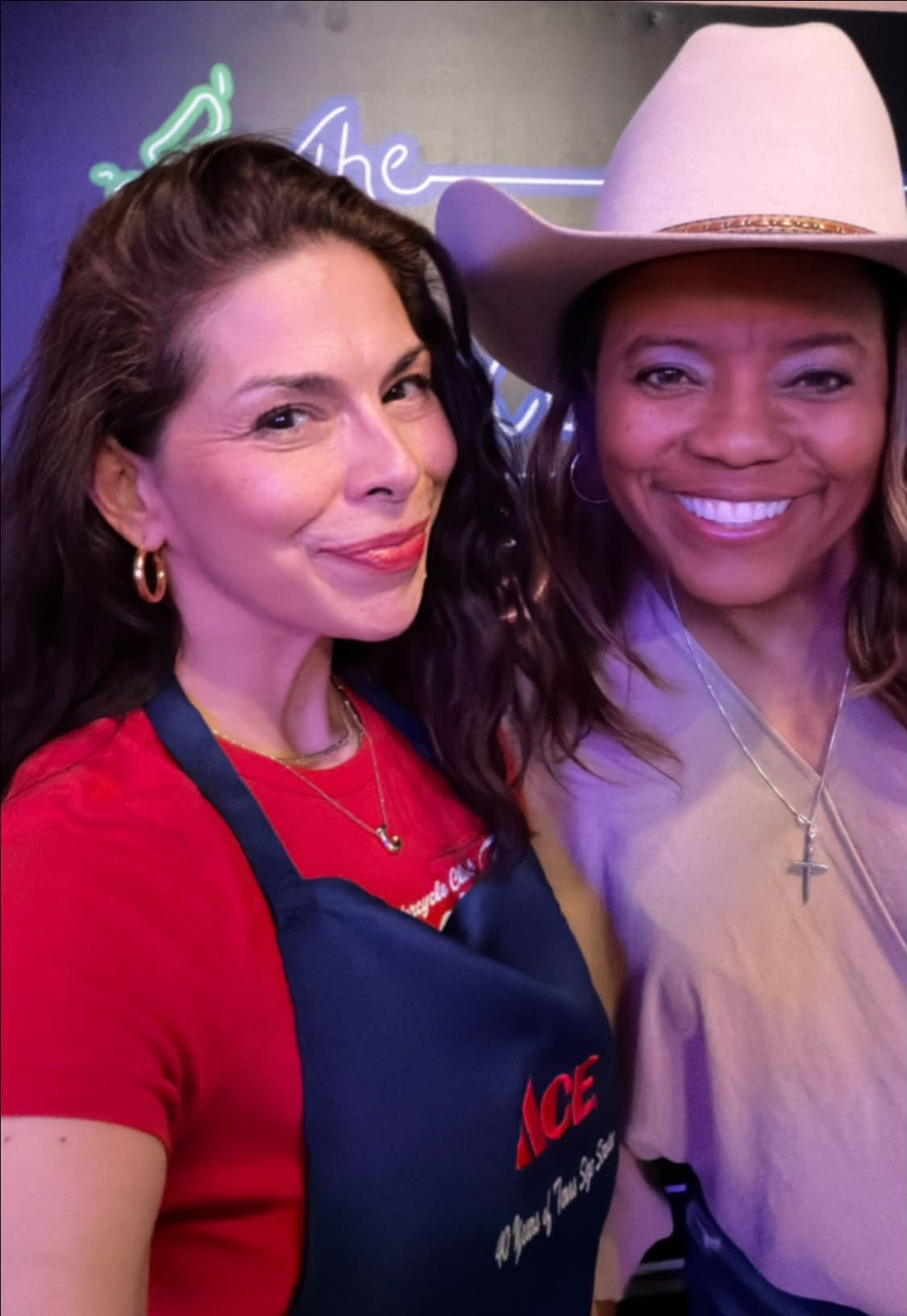 Two women smiling, one wearing a red shirt and apron, the other in a light purple top and white hat, in a neon-lit setting.