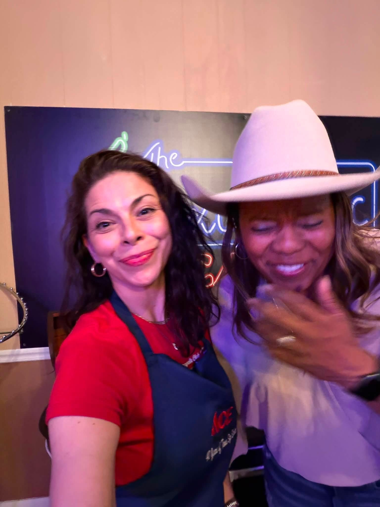 Two women smiling, one wearing a red shirt and apron, the other wearing a hat and light-colored top, in front of a dark background.