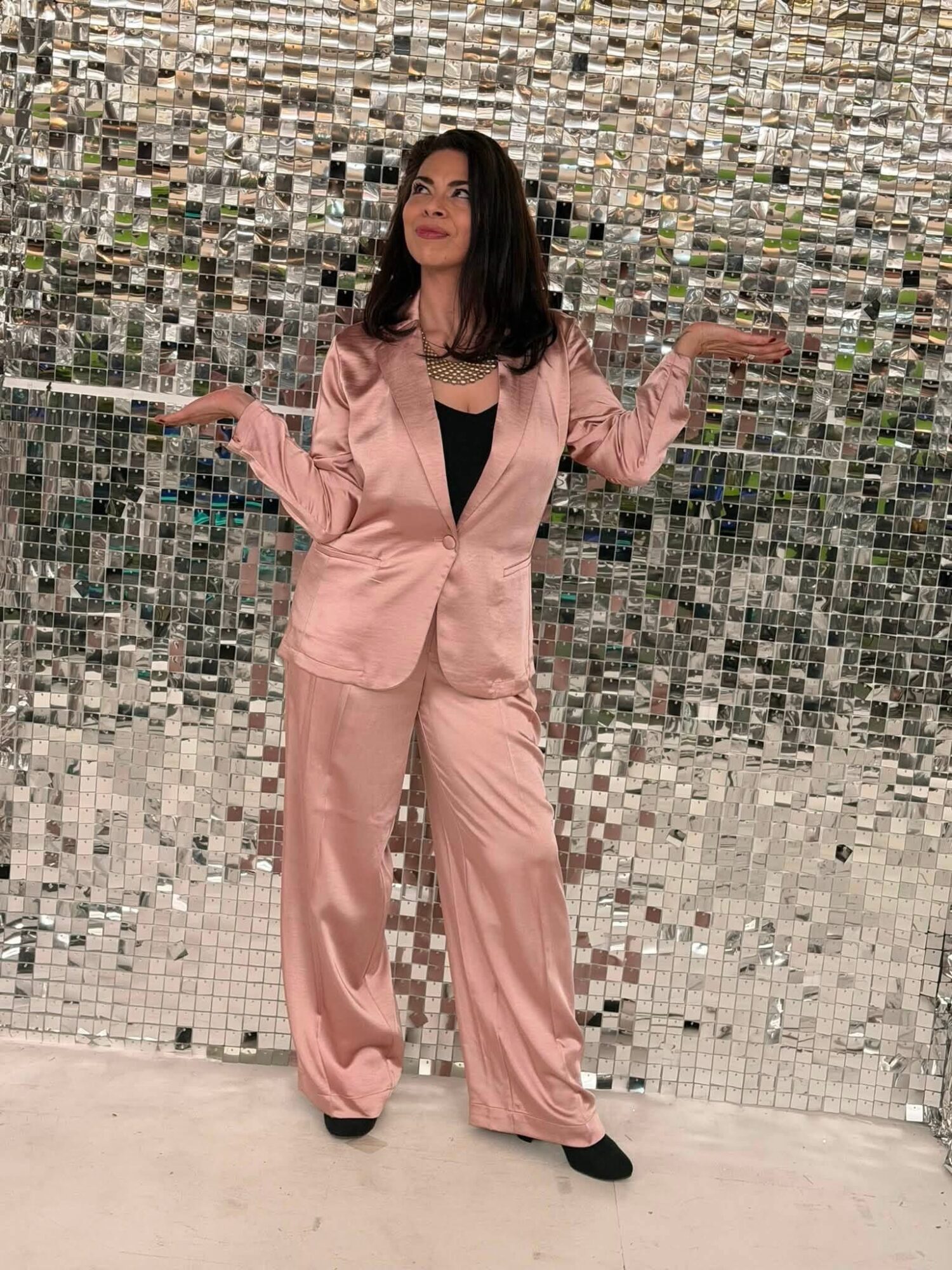 Woman in pink suit with arms raised, standing in front of a mosaic wall, looking upward.