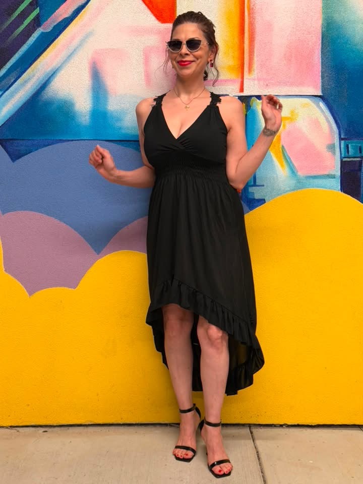 Woman in a black dress and sunglasses standing in front of a colorful mural, holding a yellow object, smiling.