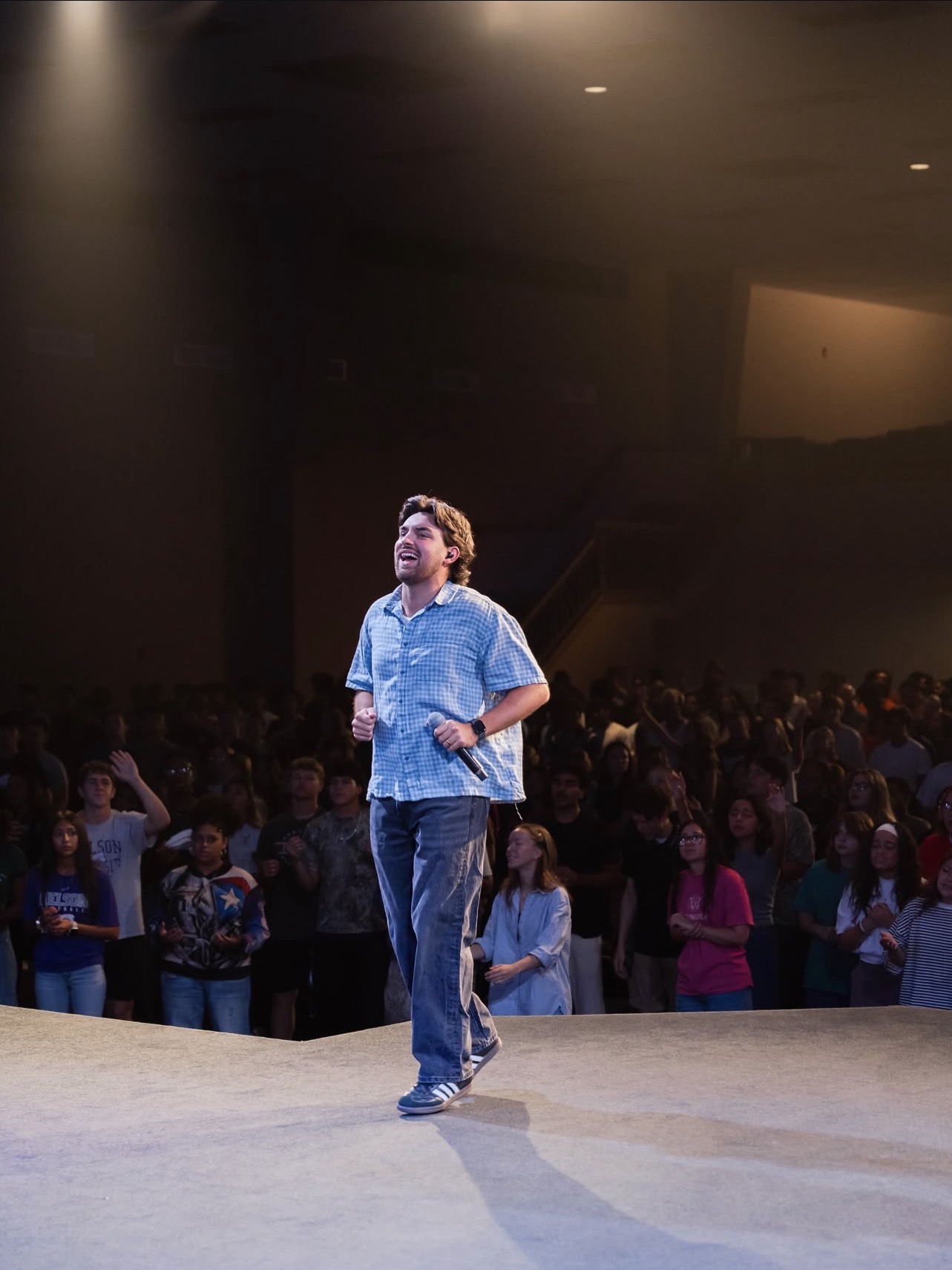 Person standing on stage in front of a large audience, wearing a denim shirt and jeans, with a spotlight overhead.